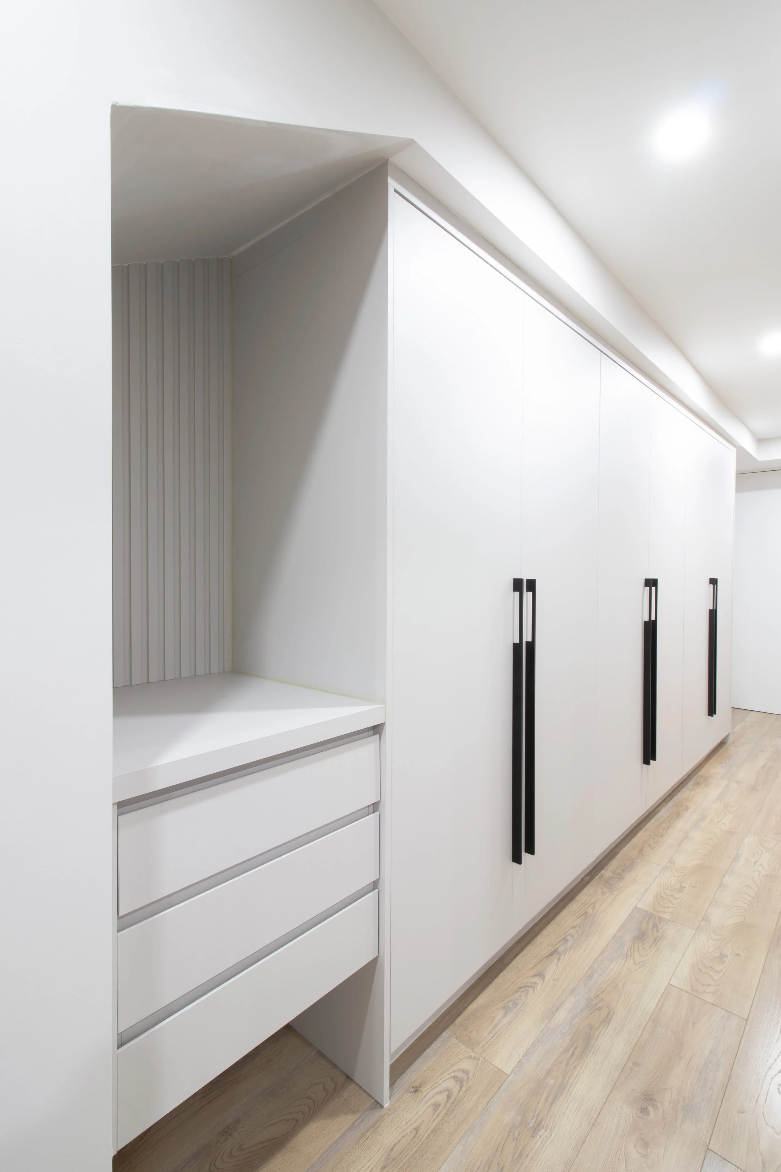 White kitchen cabinets with black handles, set against a light-colored wooden floor, with a corner shelf and drawers on the left.