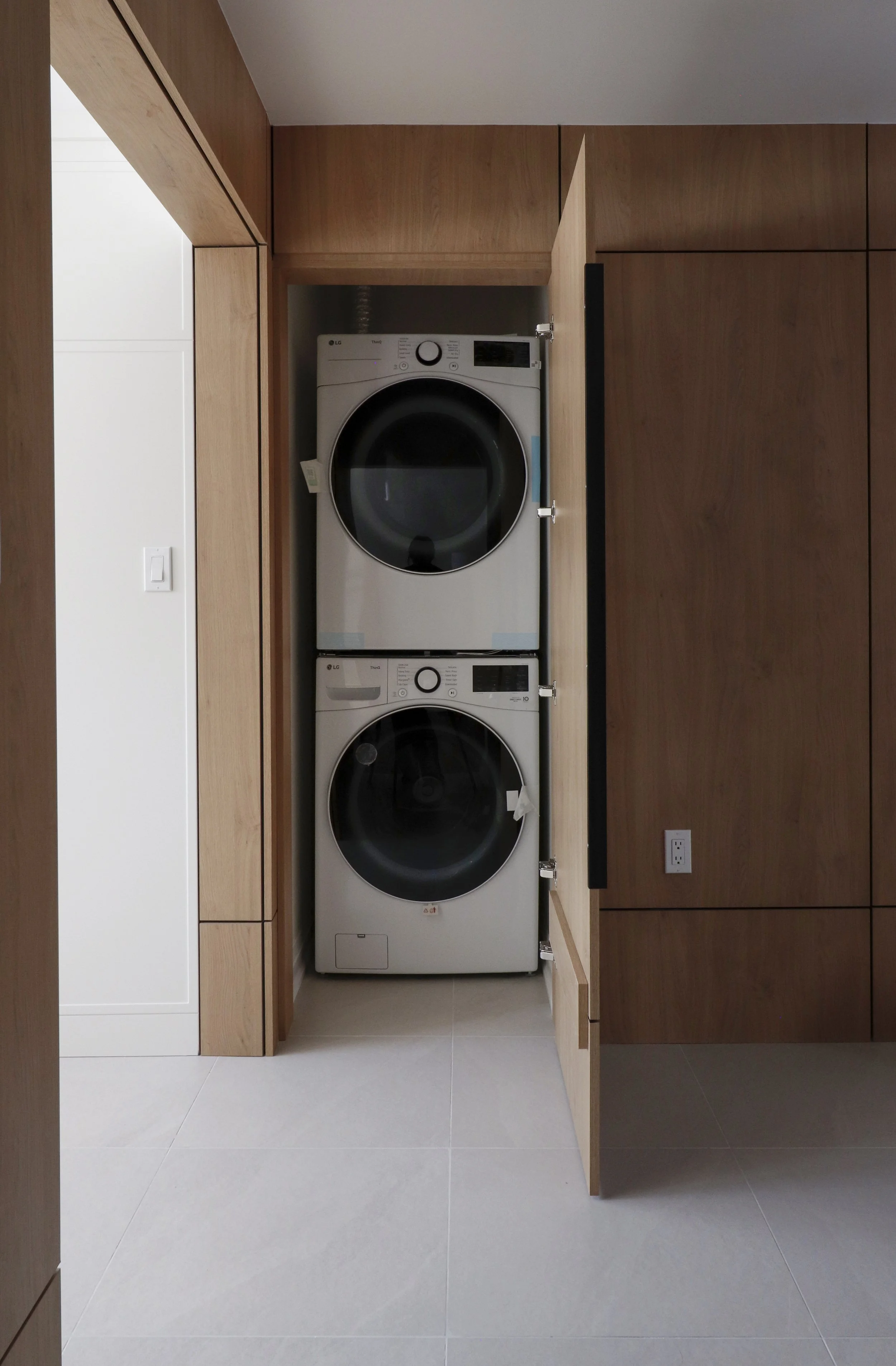 A laundry room with wooden cabinets, a stacked LG front-loading washer and dryer, white walls, tiled floor, an electrical outlet, and a sink faucet visible.