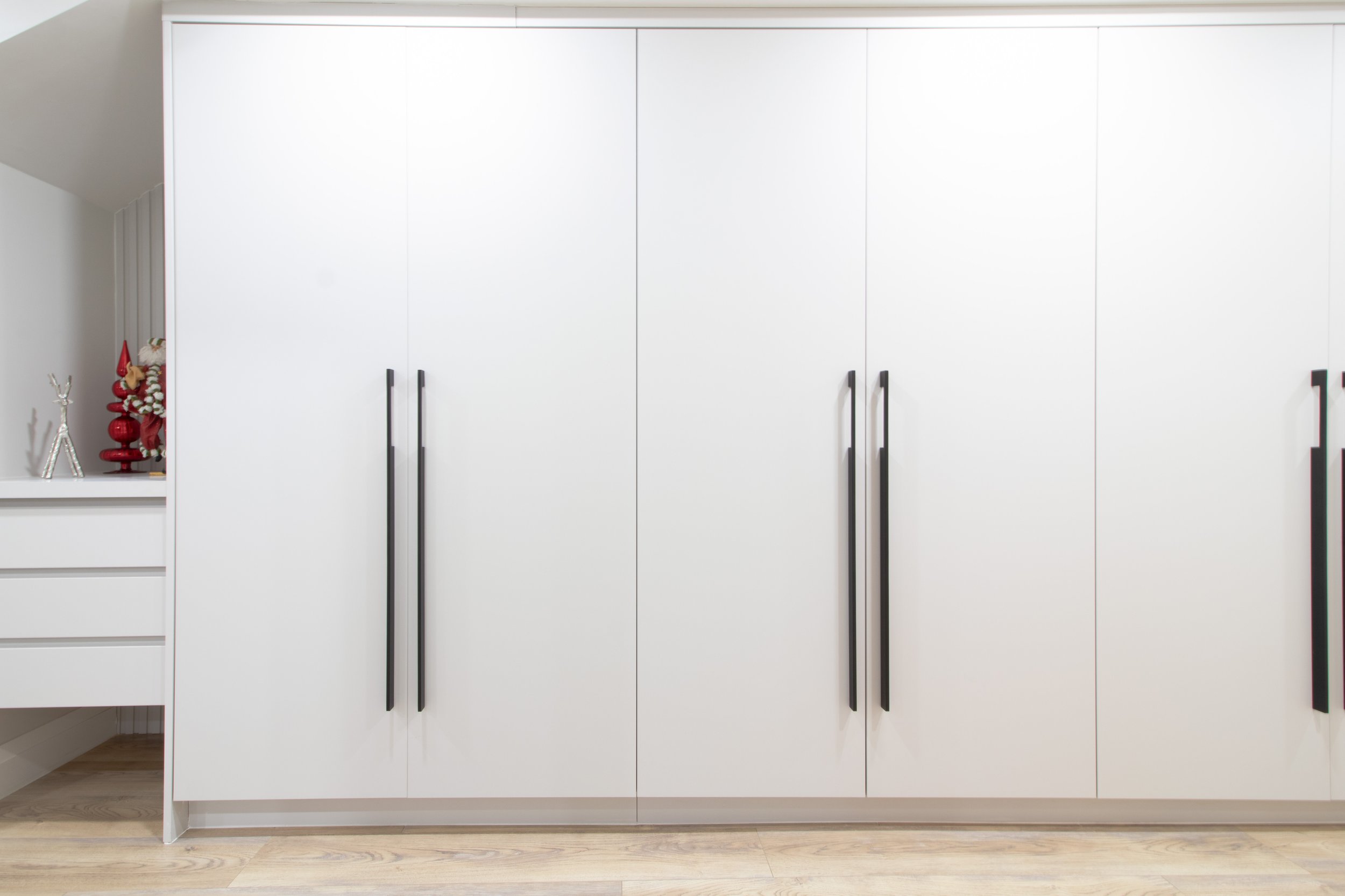 Large white closet with black handles in a modern room. To the left, Christmas decorations including a small red tree and a silver reindeer are visible.
