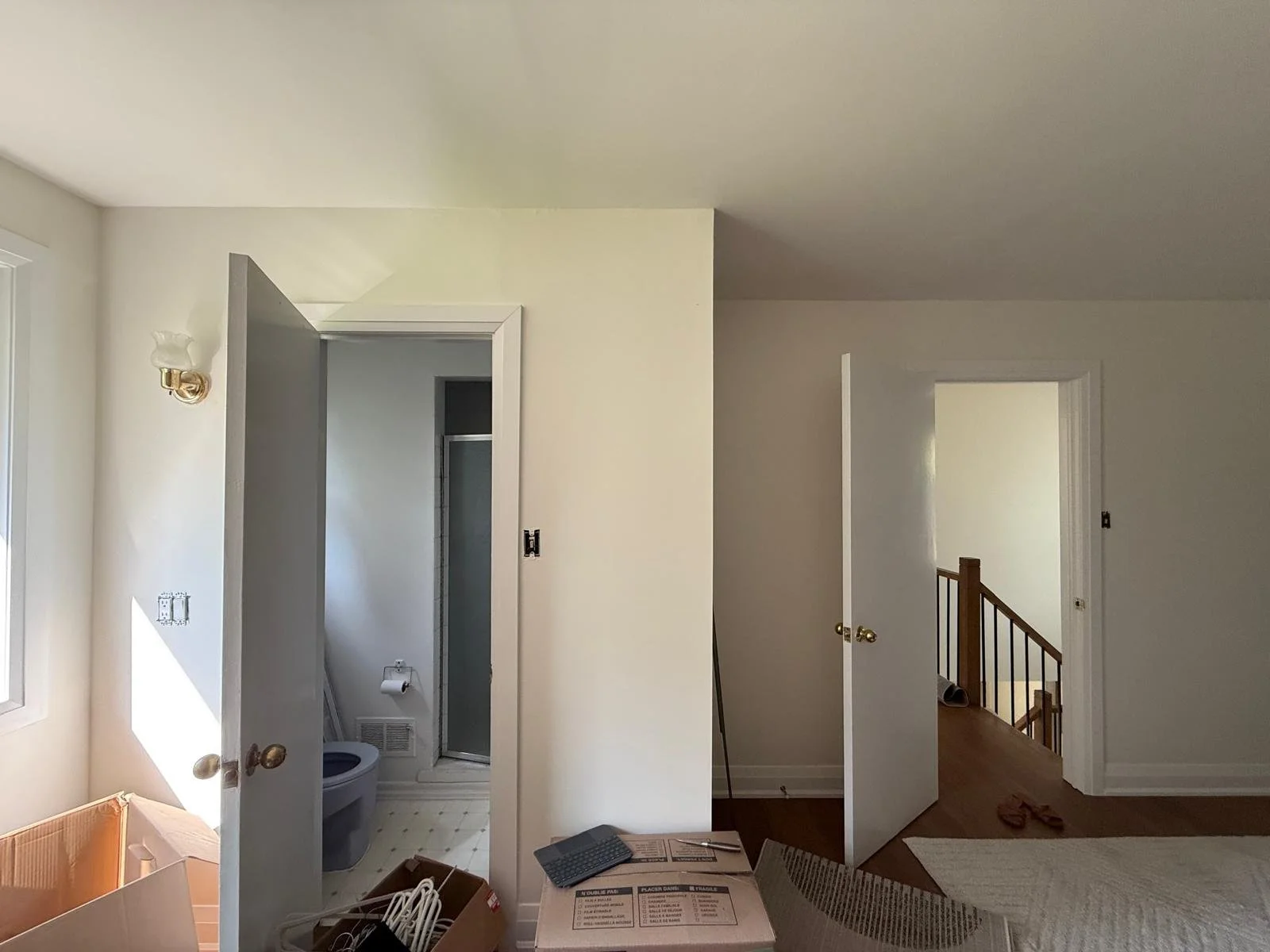 Interior view of a house with a hallway, a partially open door leading to a bathroom with a toilet and shower, and another door leading to a staircase. The space appears to be under renovation with boxes, cables, and tools on the floor.