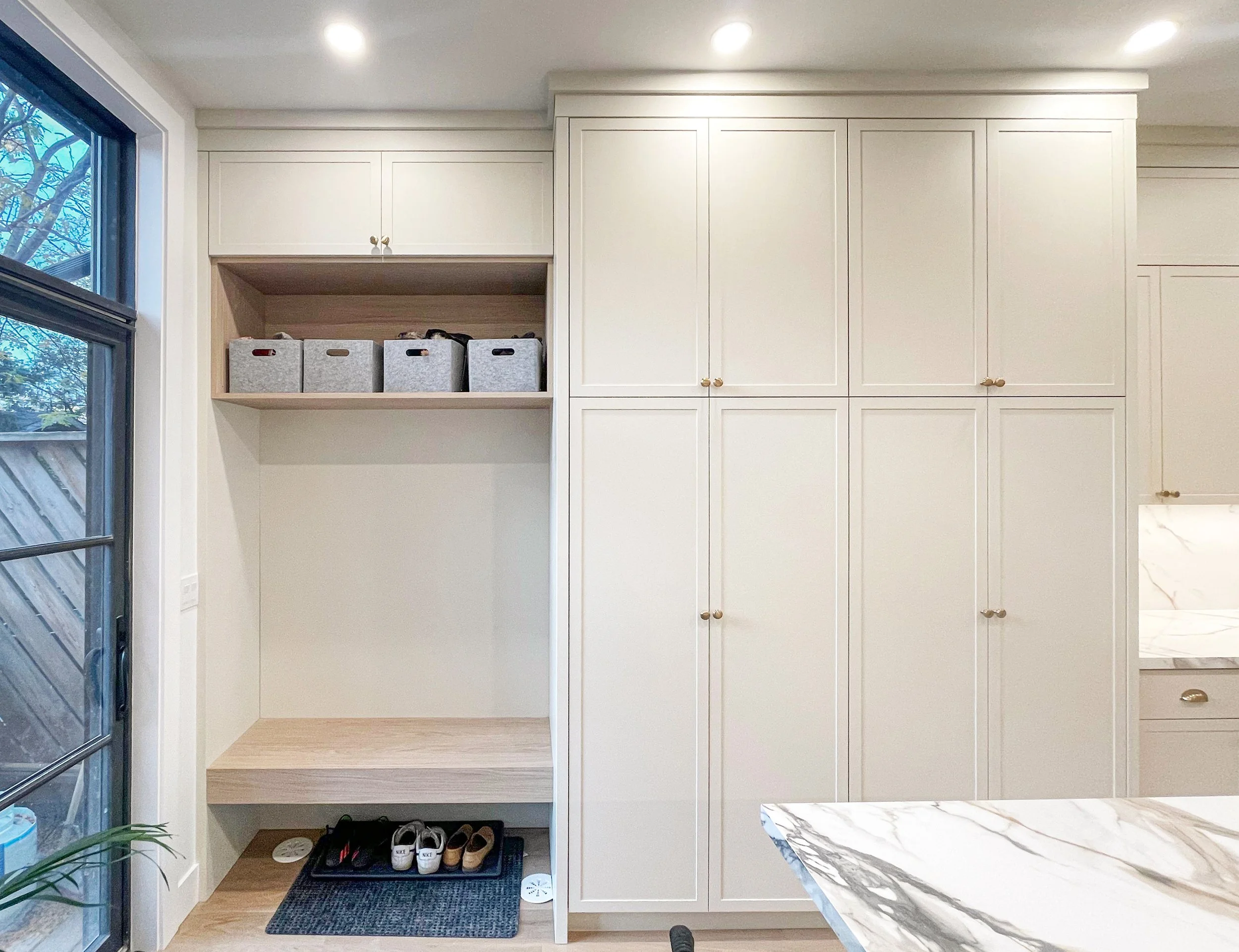 Interior view of a closet area with white cabinetry, a window on the left, and a small shoe rack at the bottom. The cabinets have gold knobs, and there are storage bins in an open shelf space. A carpeted step is in front of the shoe rack, and part of