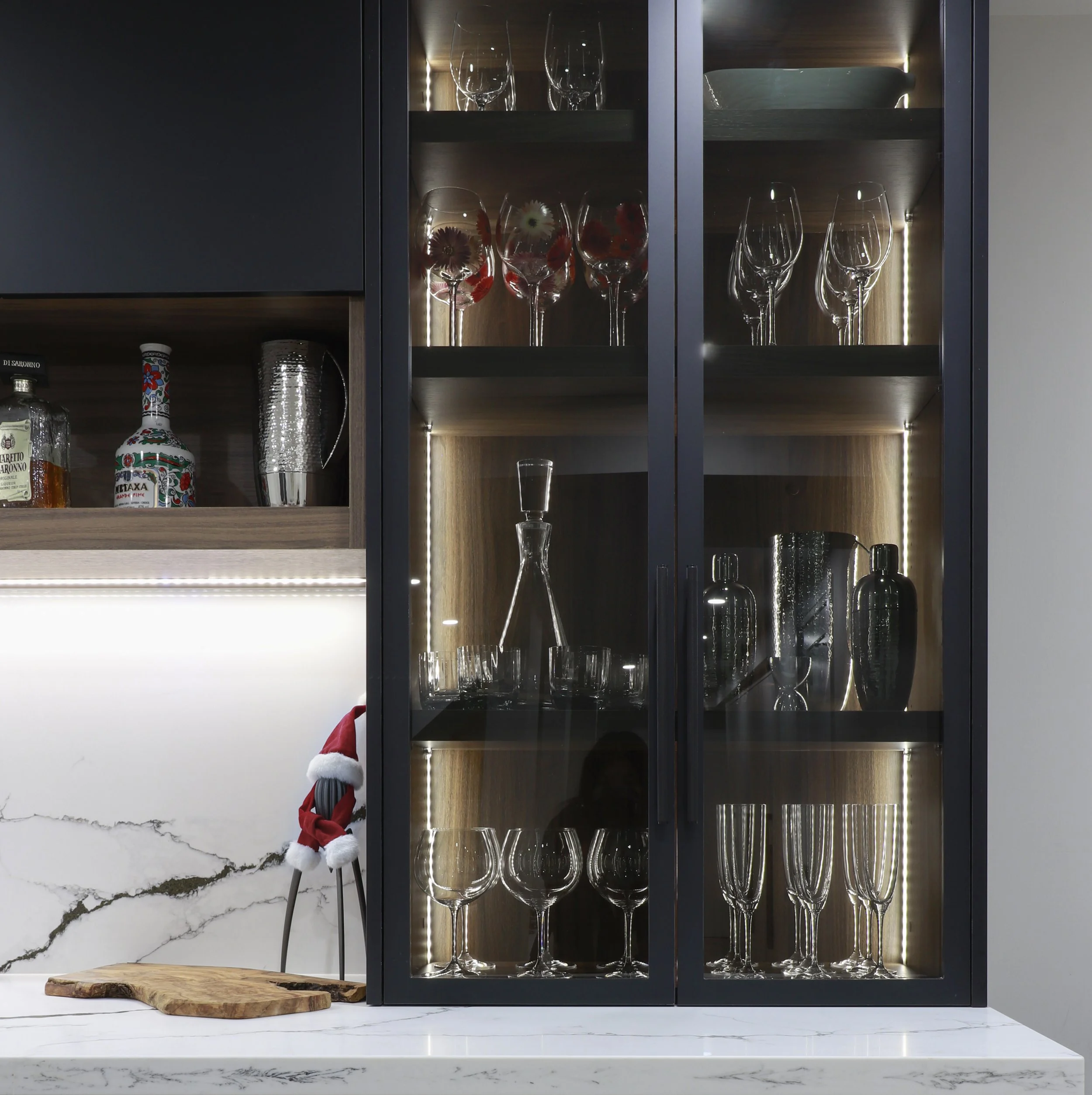 A black display cabinet with glass doors holds wine glasses on two shelves illuminated by interior lighting. Below, on a white marble countertop, there are more glasses and decorative items. A small Santa hat on a stick and a wooden cutting board are