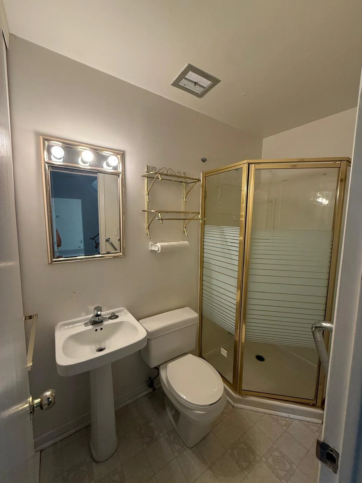 Small bathroom with a pedestal sink, toilet, and shower enclosure with gold trim. Wall-mounted mirror with four lights, metal shelving above the toilet, and a paper towel holder. Gray tile floor and light-colored walls.
