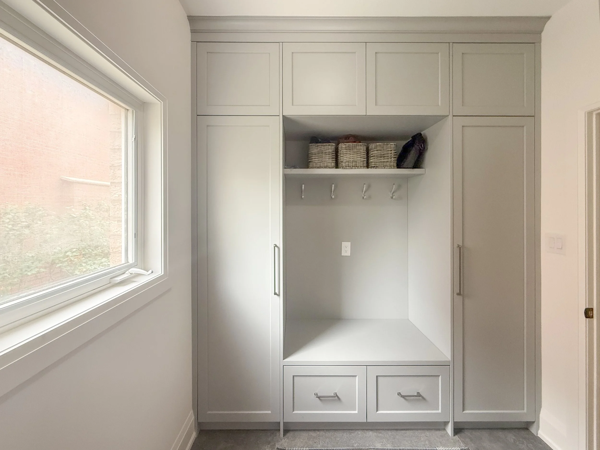 Empty white built-in closet with upper cabinets, middle open shelves with woven baskets and a black bag, and lower closed cabinets, next to a large window in a bright room.
