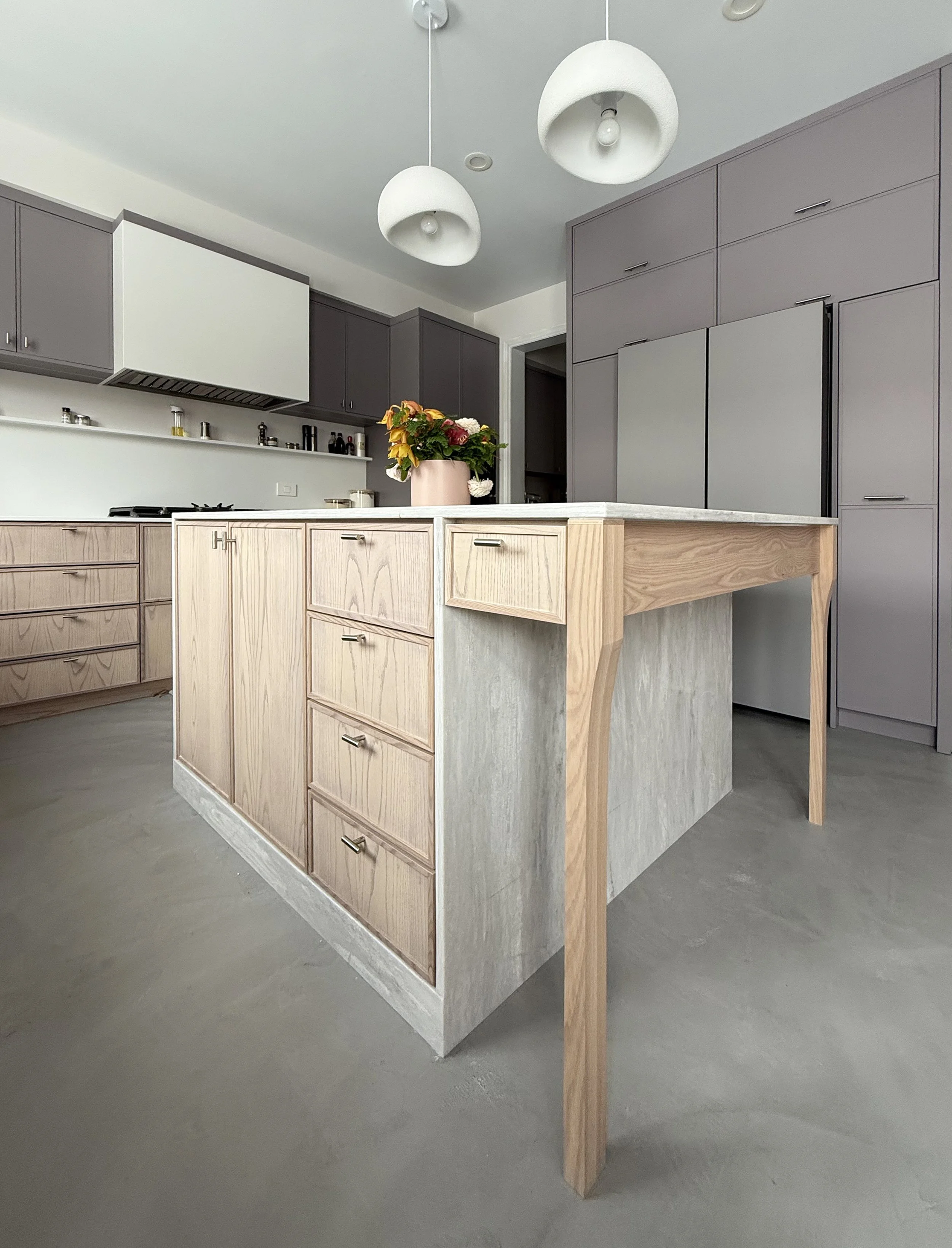 Modern kitchen with light wood island, gray cabinets, white countertops, a flower arrangement in a pink vase, and two white pendant lights.