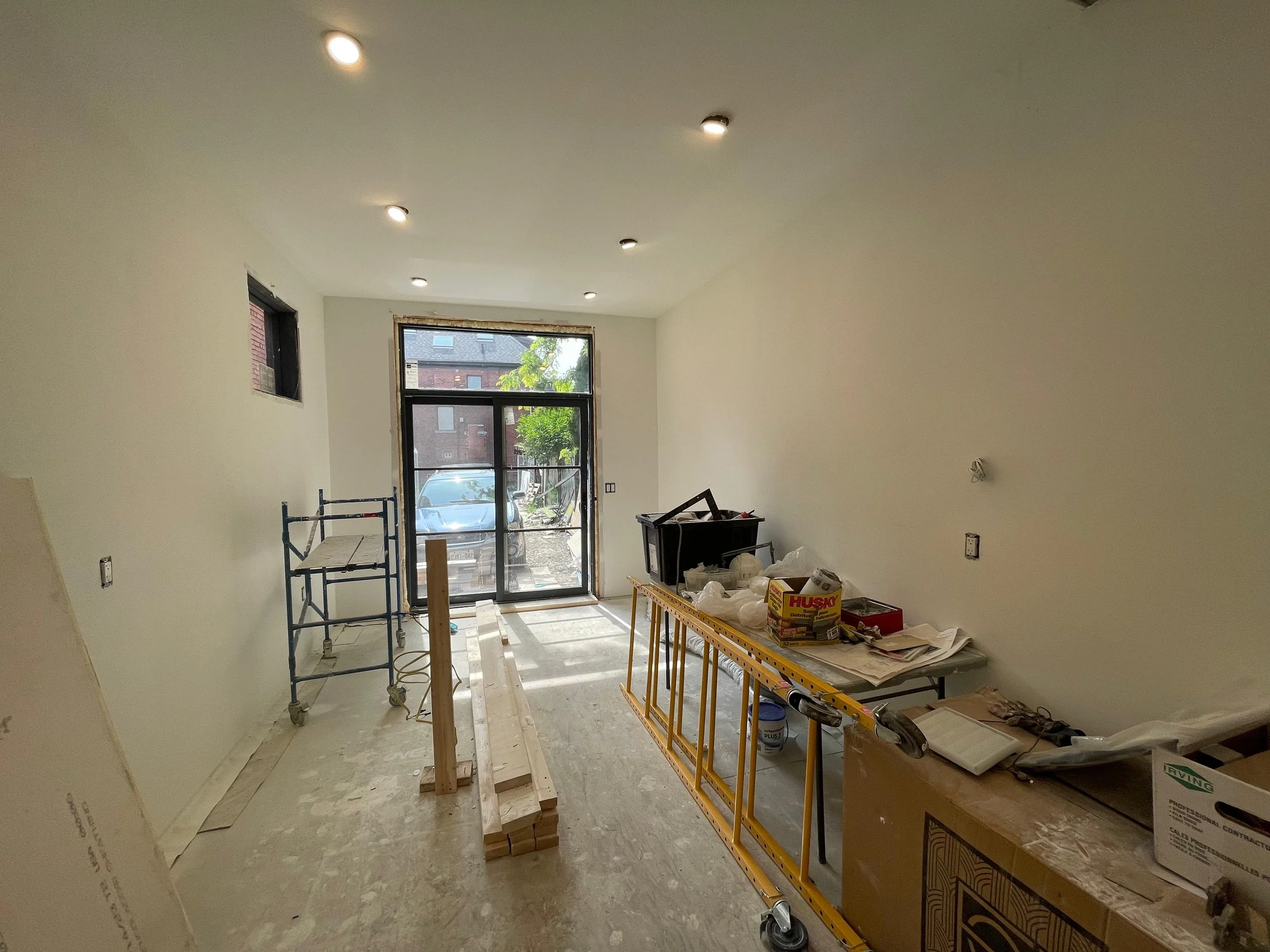 Interior of a room under construction or renovation with construction materials, a scaffold, a work table, and construction supplies near the window showing a parked car outside.