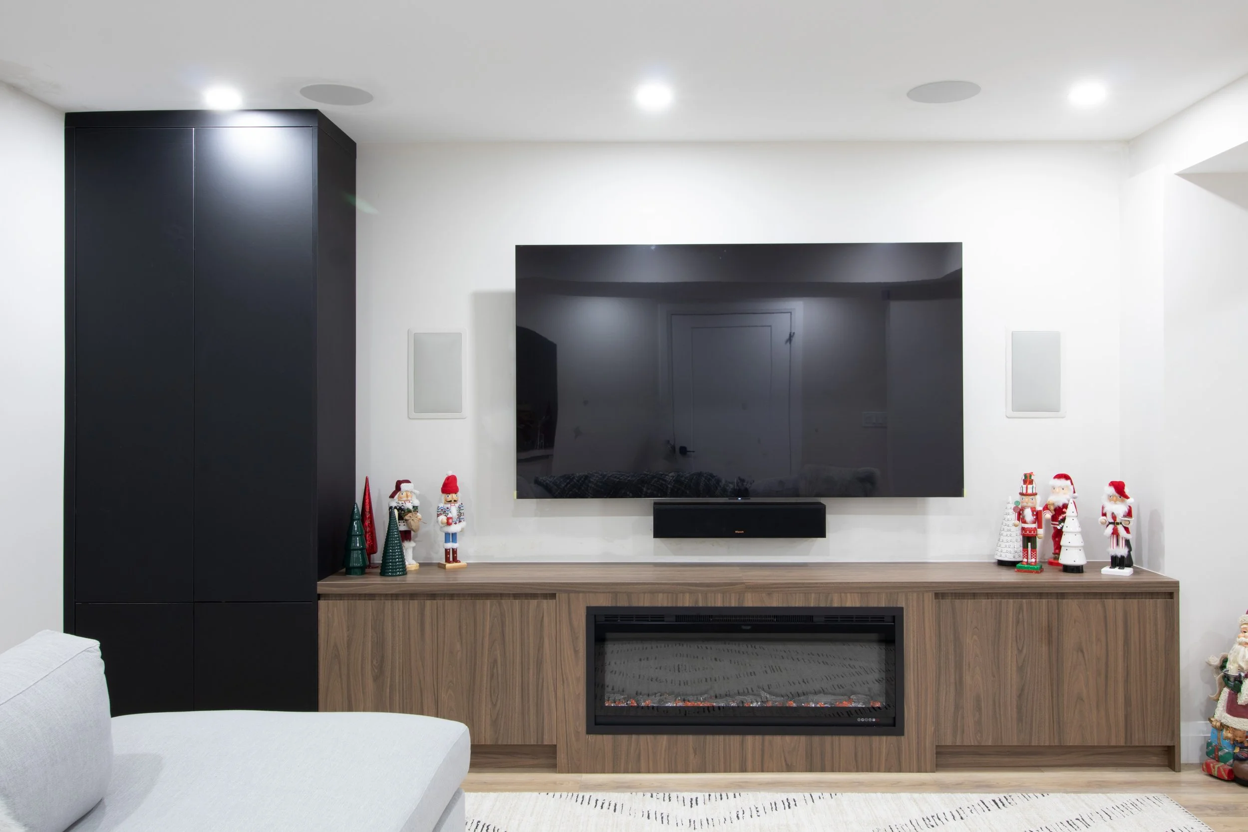 Living room with a wall-mounted flat-screen TV, black cabinet, wood fireplace, holiday decorations including nutcracker figures, Christmas trees, and Santa Claus figurines