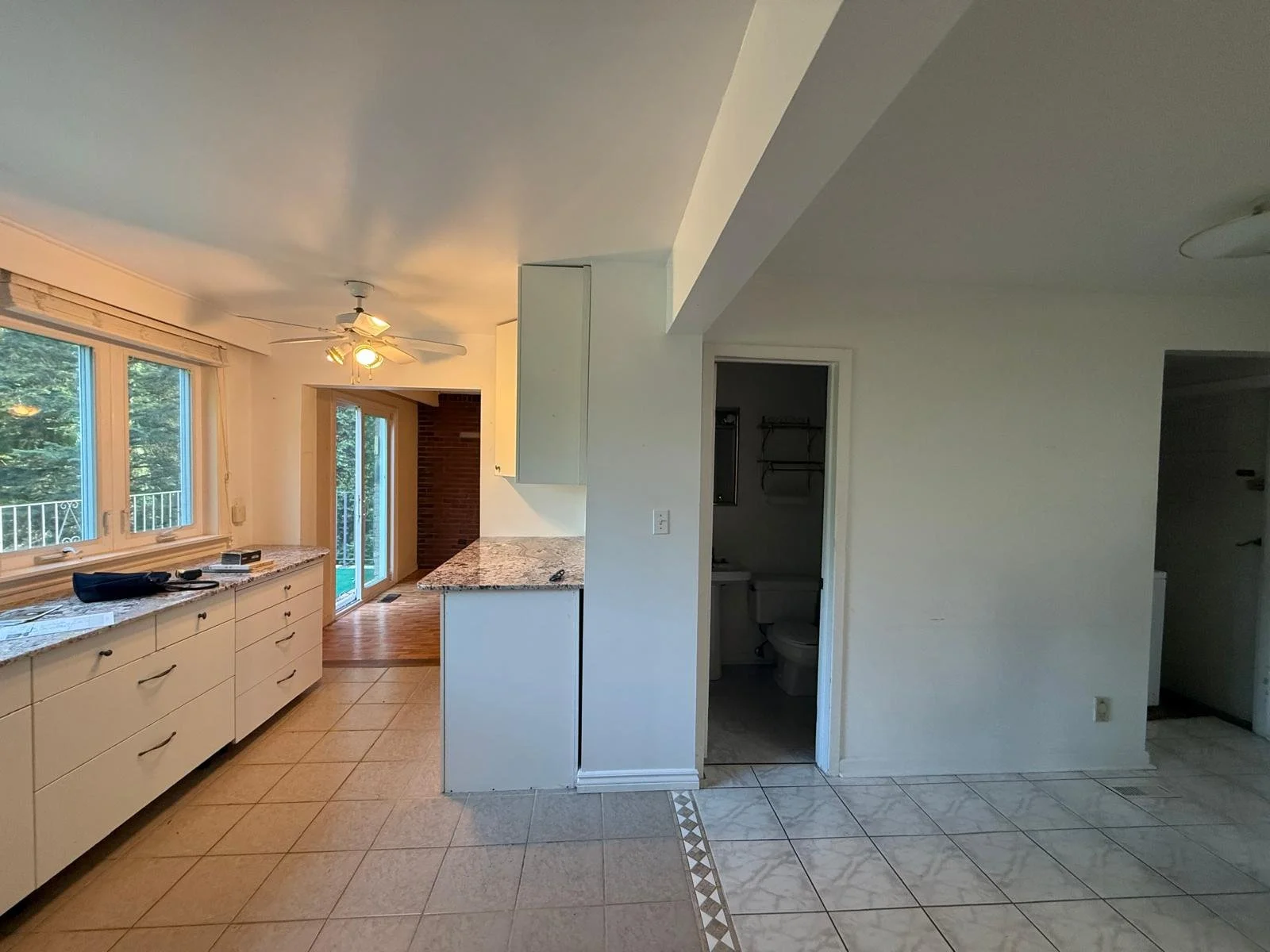 Interior view of a kitchen with white cabinetry, granite countertops, large windows, a ceiling fan, and a sliding glass door leading to an outside area; adjacent to a small bathroom or closet with a toilet and shelves.
