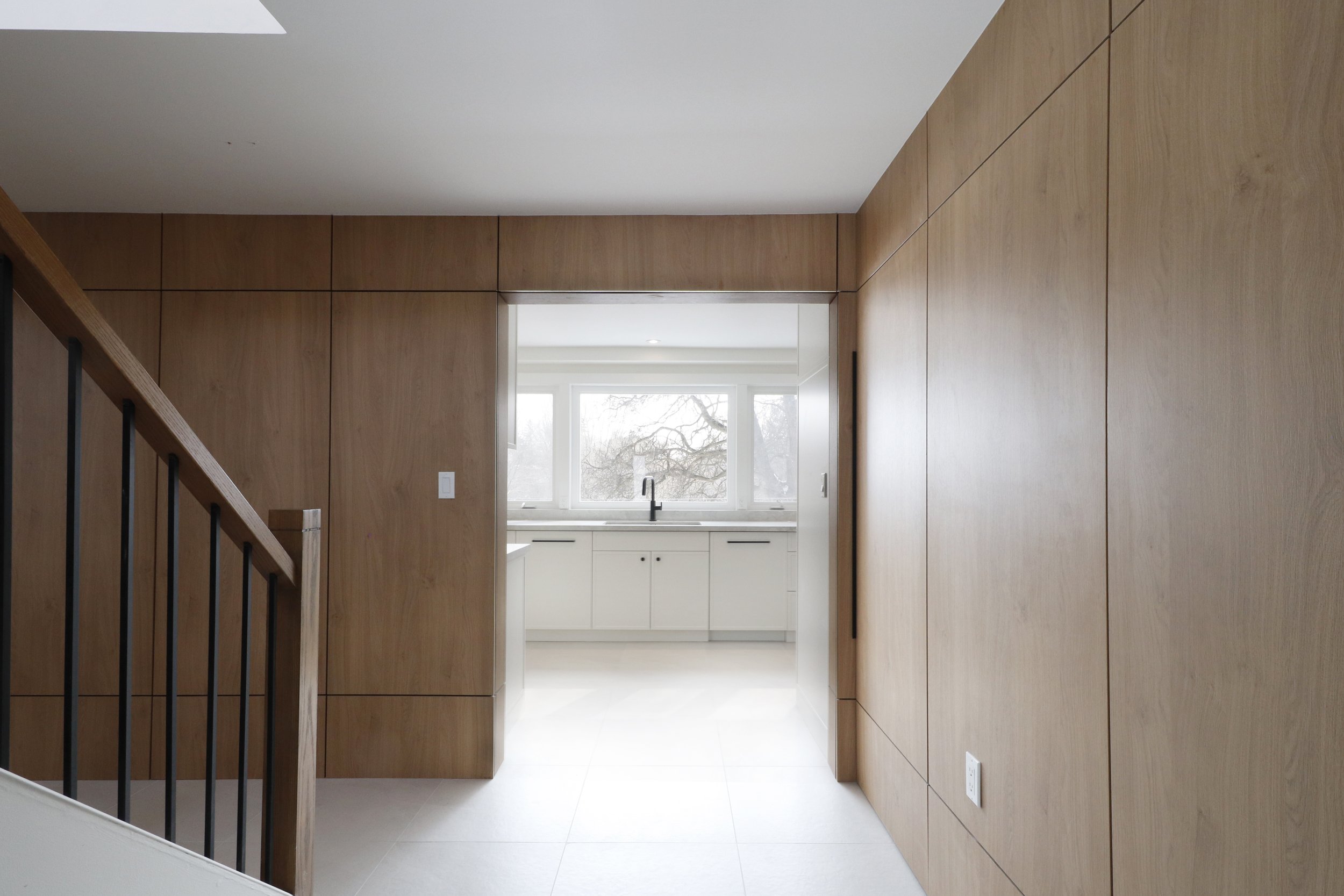 A modern kitchen with white cabinets, a large window showing trees outside, and light-colored flooring. The photo is taken from bottom of stairs with wood railing.