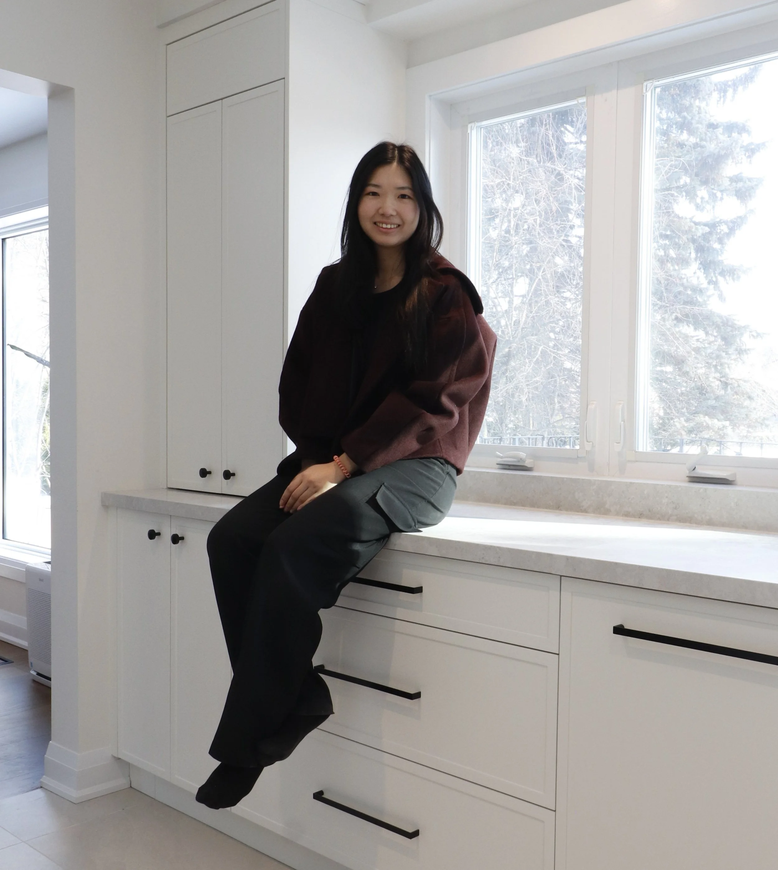 A young woman with long black hair is sitting on a kitchen counter near a large window, smiling at the camera. She is wearing a dark top and gray pants, with a red bracelet on her right wrist. The kitchen has white cabinets, a white countertop, and a view of snow-covered trees outside.