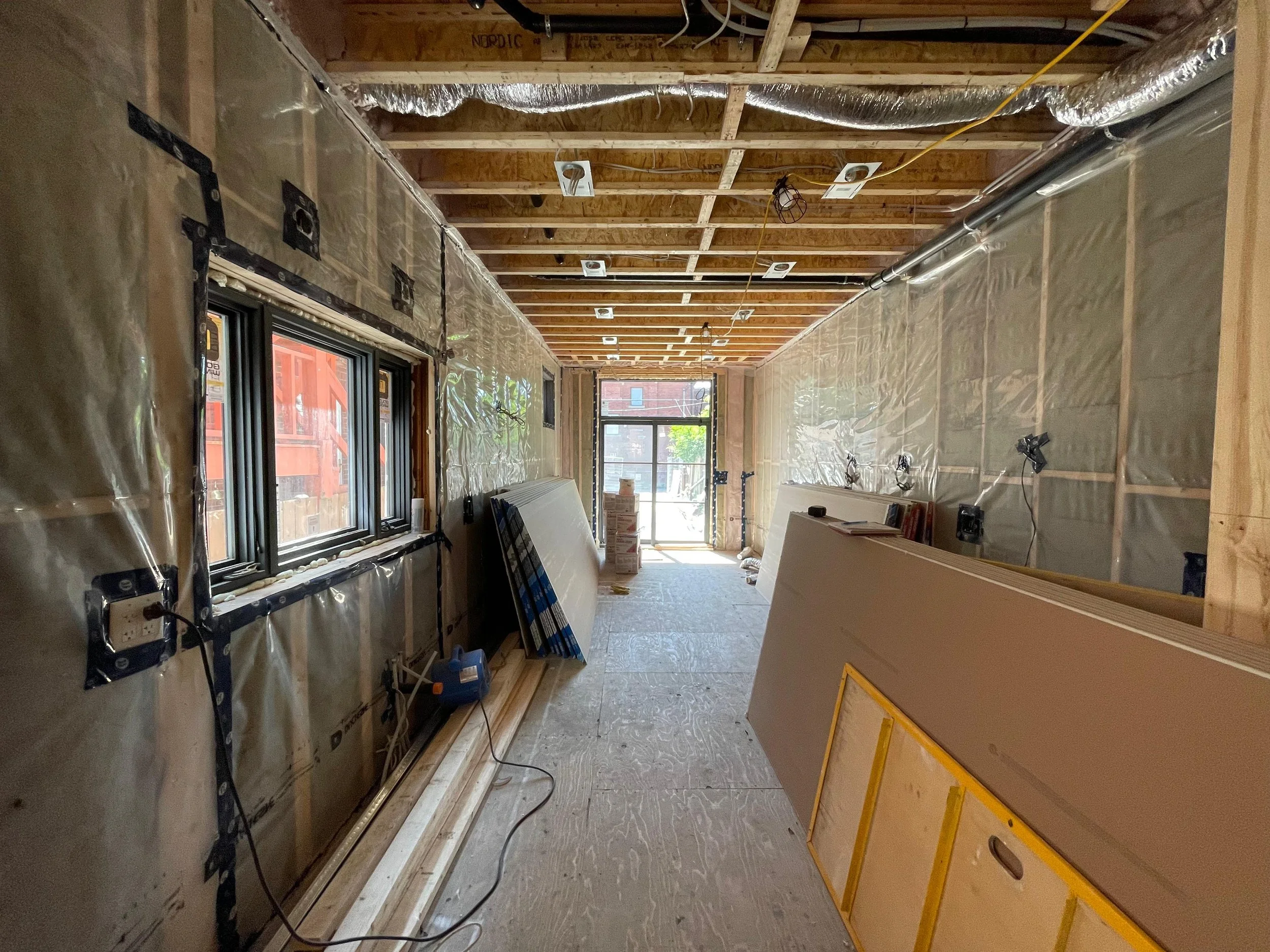 Interior of a room under construction with exposed insulation, wiring, and unfinished wooden framing, large windows at the far end, and construction materials and tools scattered on the floor.
