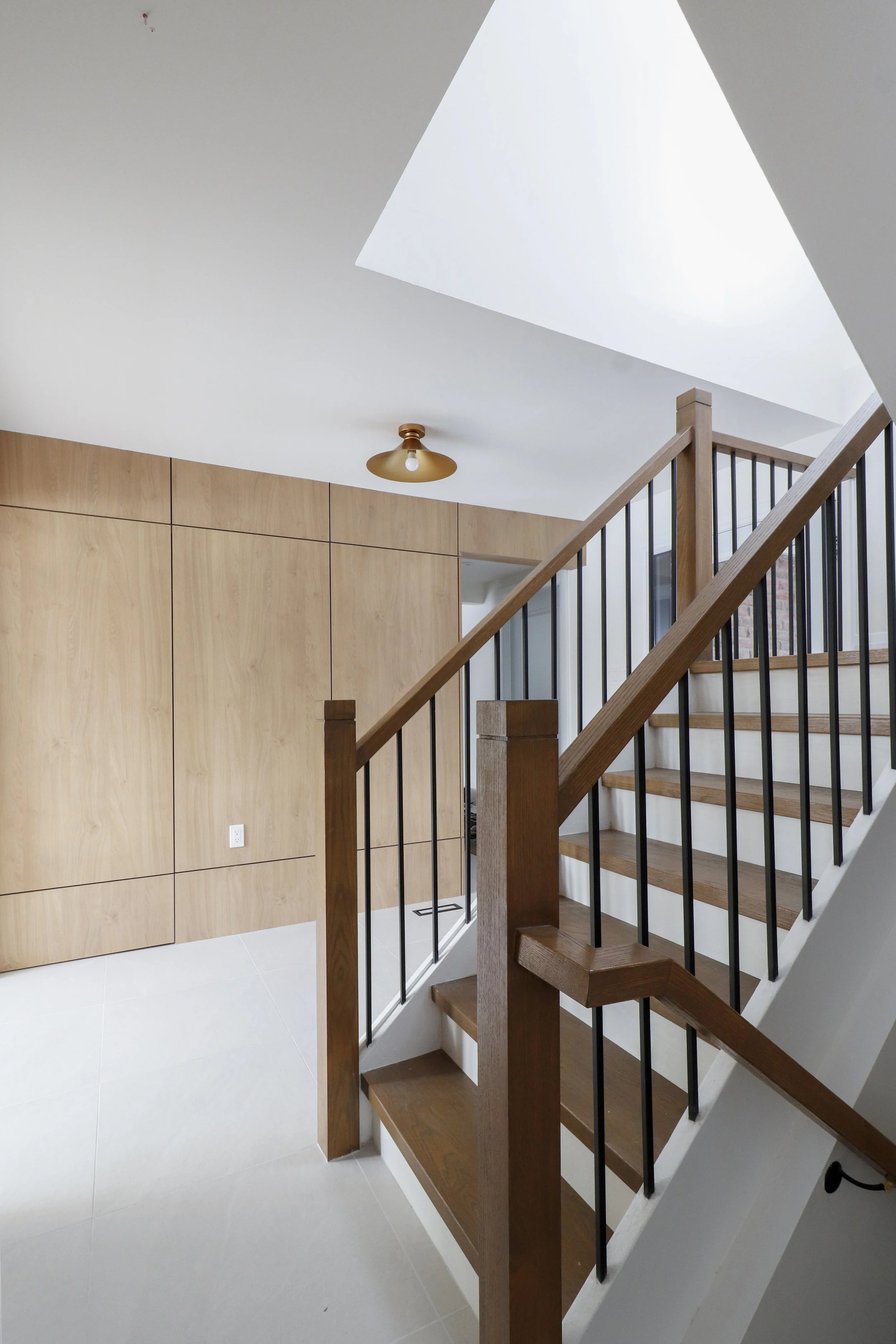 Modern staircase with wooden steps and black metal railing, next to a light wood-paneled wall, in a bright interior with a large skylight and minimal decor.