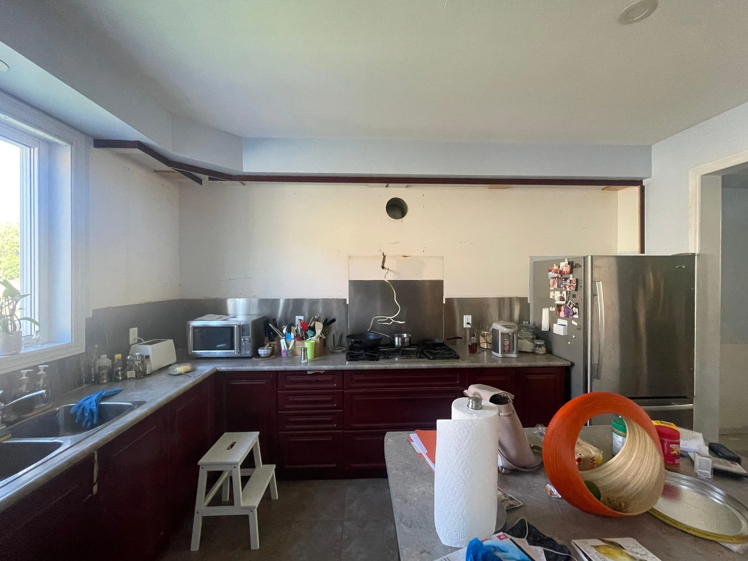 A cluttered kitchen with red cabinets, a granite countertop, a window, a microwave, a stainless steel refrigerator covered with photos, and various kitchen items and paper towels on the table and counter.