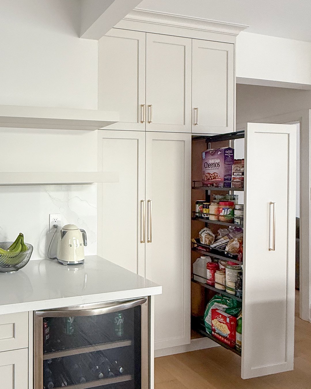 Open pantry in white kitchen with shelves full of snacks and food items, cupboards above, countertop with a blender and bananas, wine fridge below, and part of a doorway on the right.