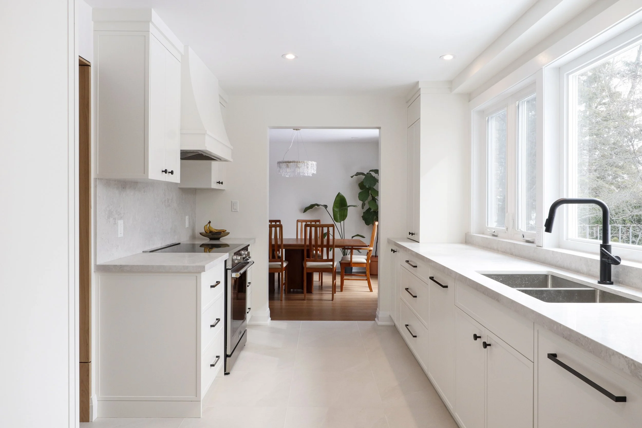 Bright white kitchen with large windows, marble countertops, black cabinet handles, stainless steel stove, and a view of the dining area with wooden table and chairs, and large green plants.
