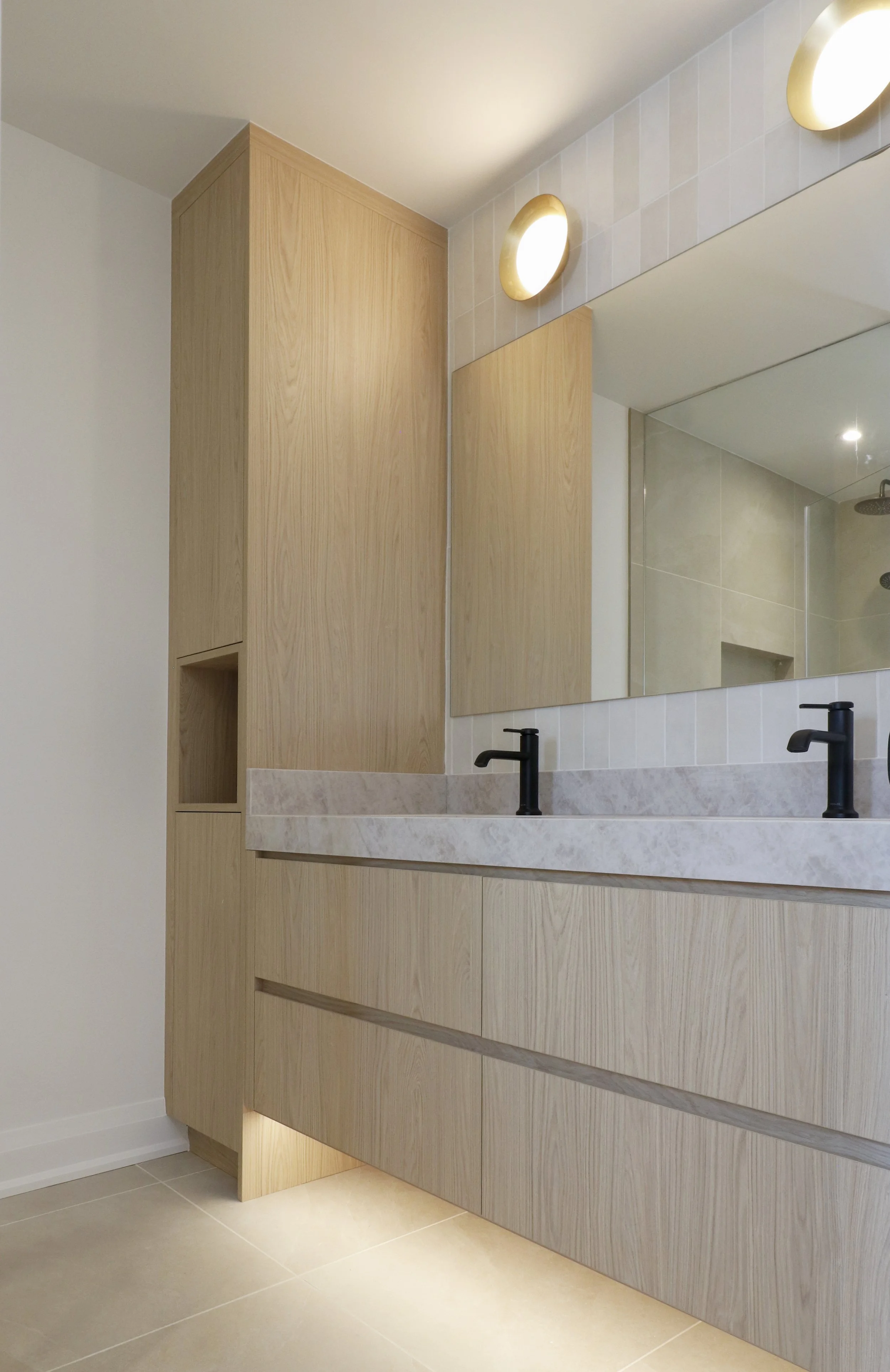 Modern bathroom vanity with light wood cabinets, a marble countertop, black faucets, a large mirror, and stylish wall-mounted light fixtures.