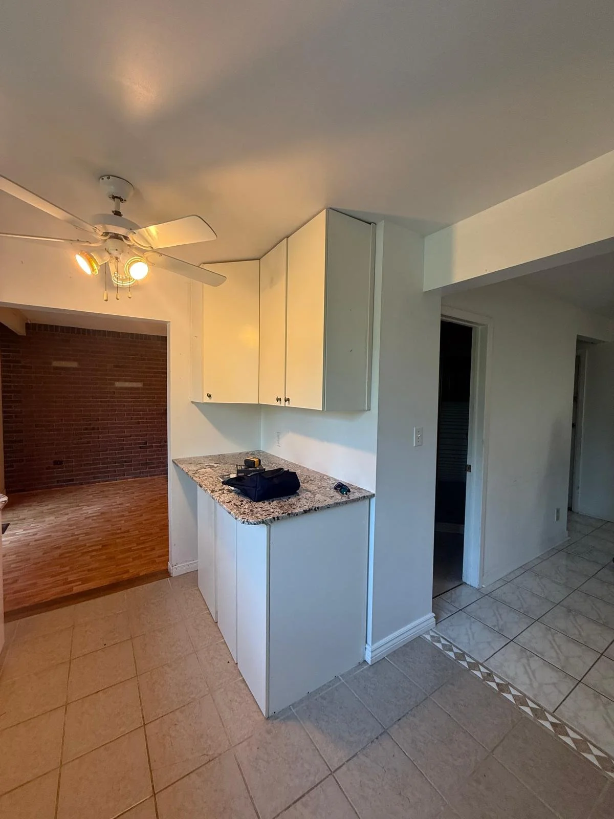 The image depicts a kitchen area with white cabinets, a granite countertop, and a tiled floor. There is a ceiling fan with lights. On the countertop, there is a black bag, a tape measure, and some small items. To the left, there is a room with a bric