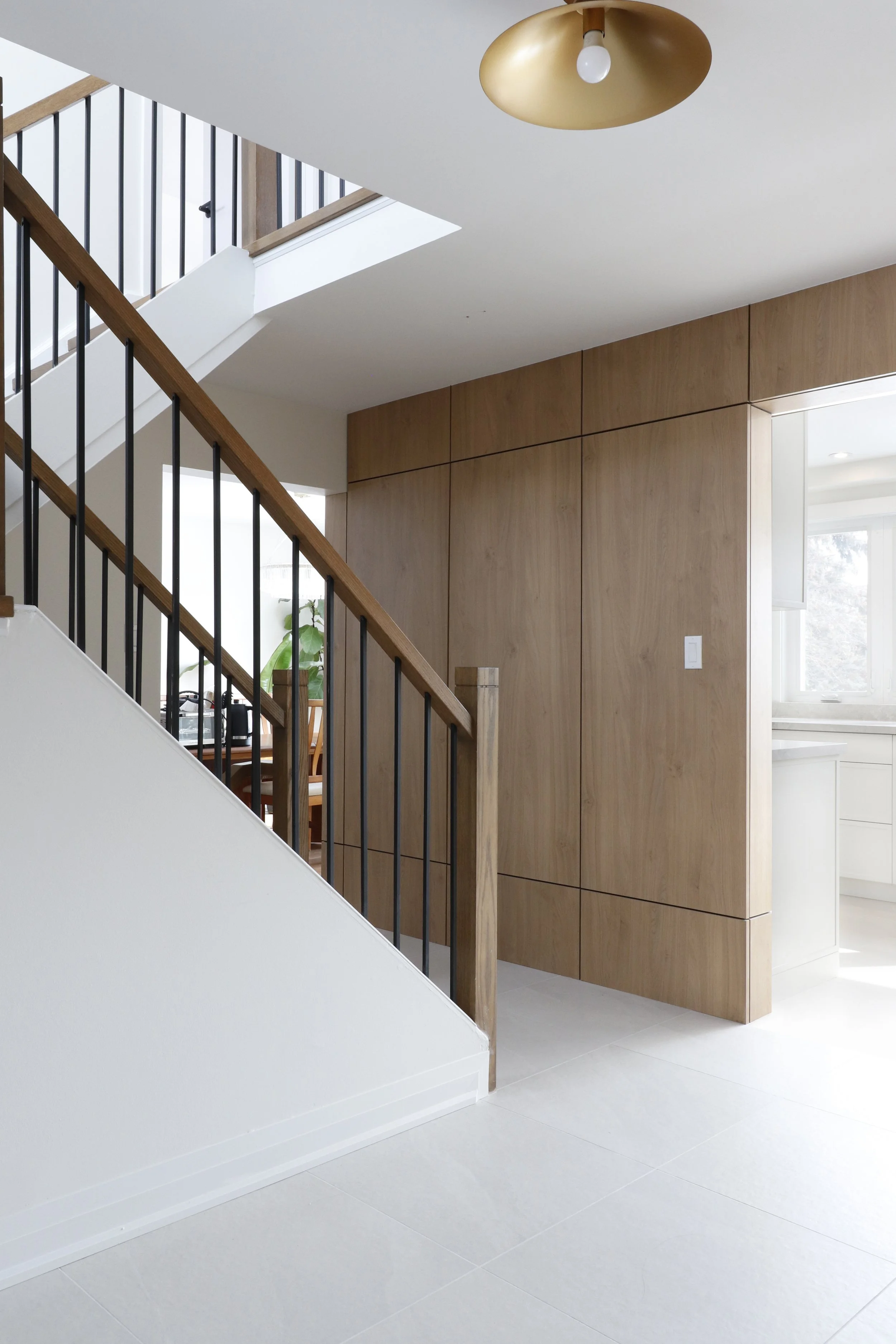 Interior view of a modern staircase area with wooden handrails and black metal balusters, adjacent to a wall with wood paneling and a white kitchen in the background, under a ceiling with a gold-colored light fixture.