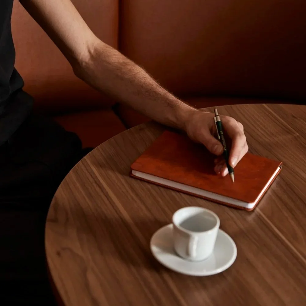 A person's arm holding a pen over an orange notebook on a wooden table, with a white cup of coffee on a saucer nearby.