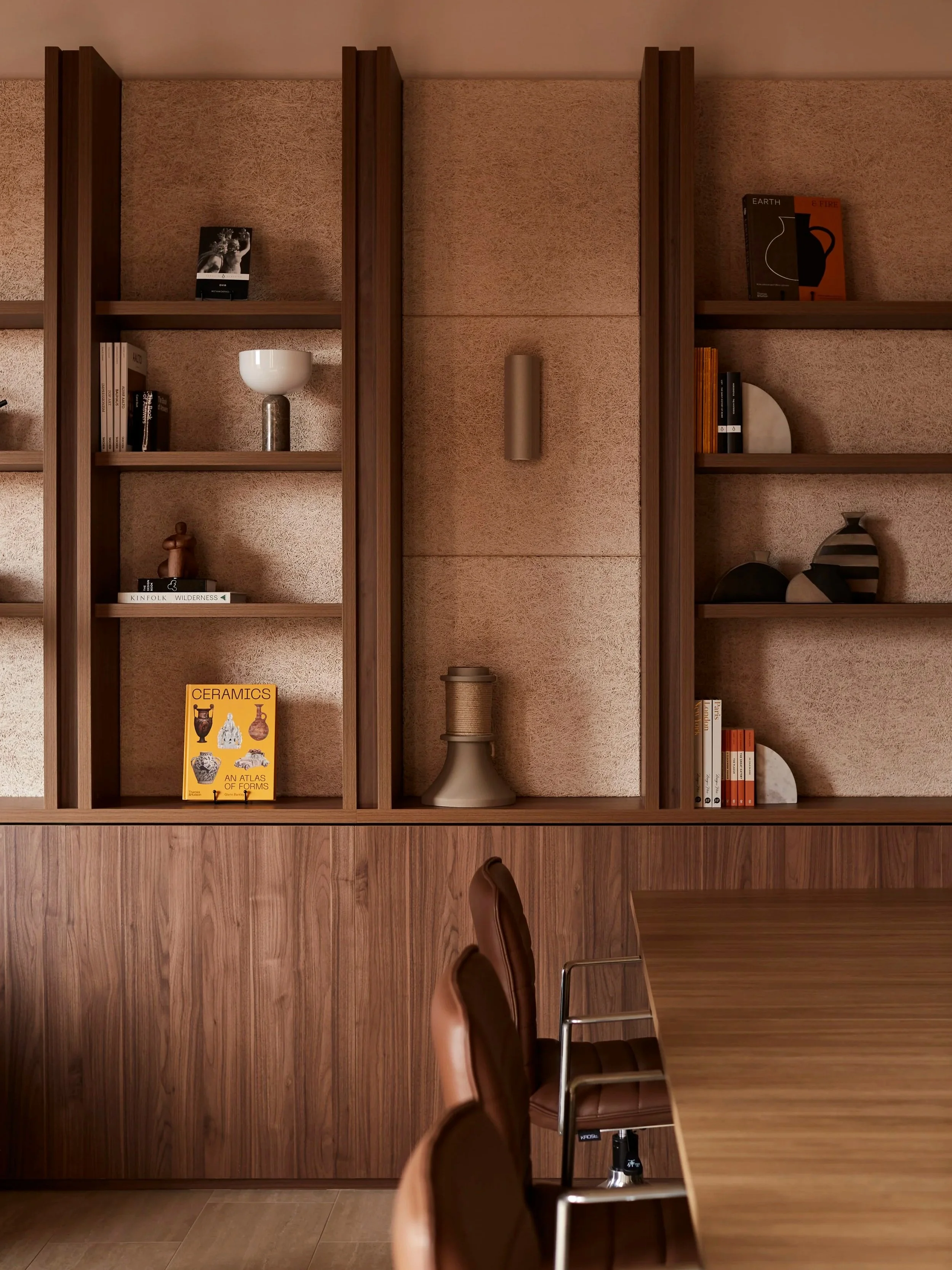 Wooden bookshelf with decorative objects and books, a continuation of a wood-paneled wall, a wooden table, and brown leather chairs in a modern interior.