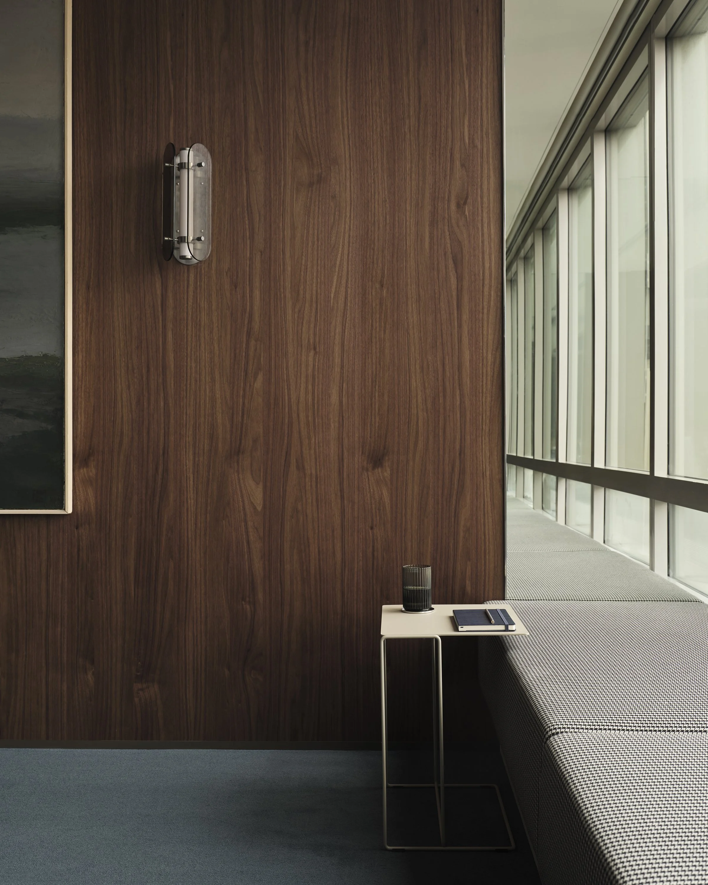 Interior space with a wood-paneled wall, a side table with a glass of dark liquid and a notebook, a window with natural light, and a cushioned seating area.