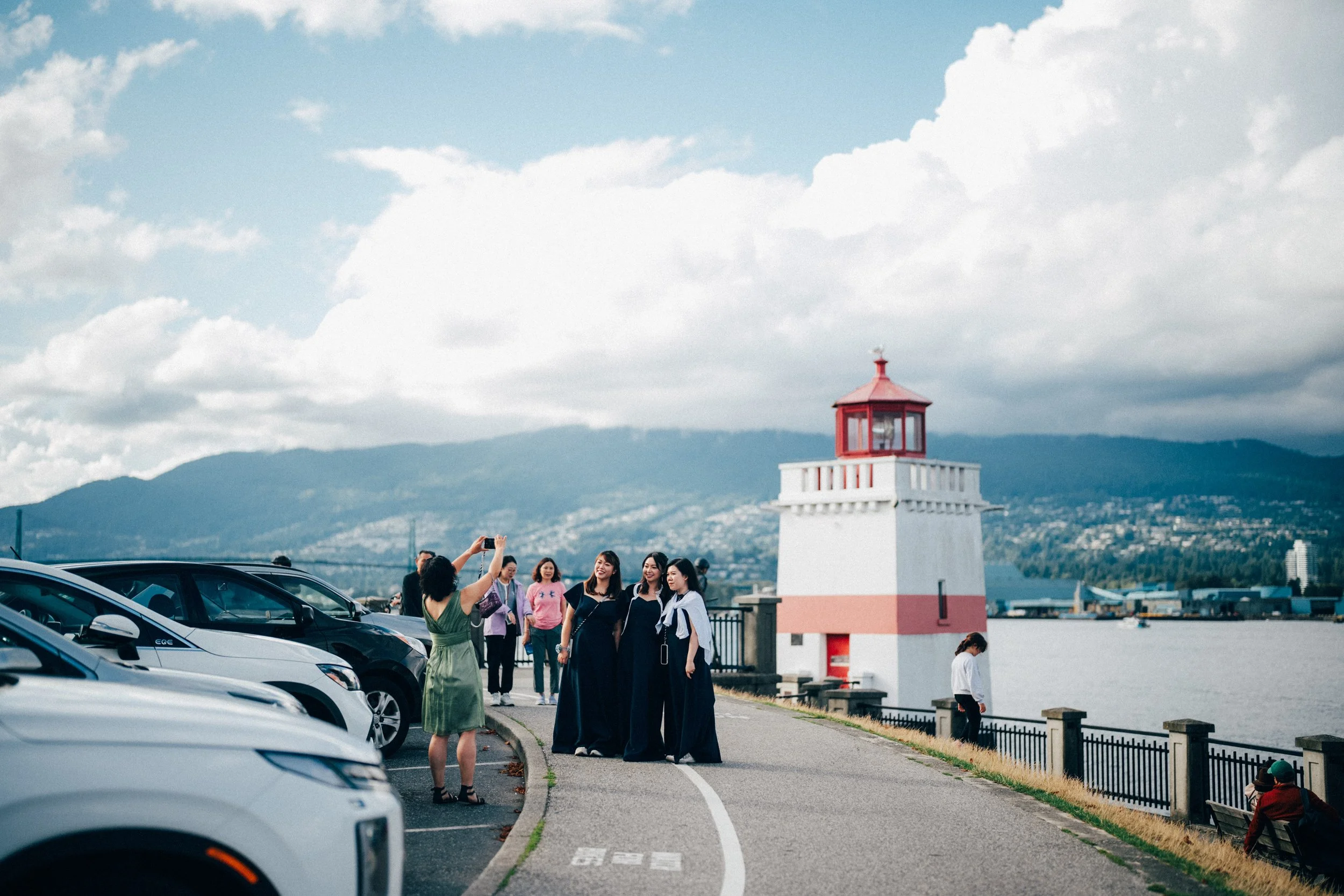 Wedding guests posing near parked cars and a red-and-white waterfront tower.