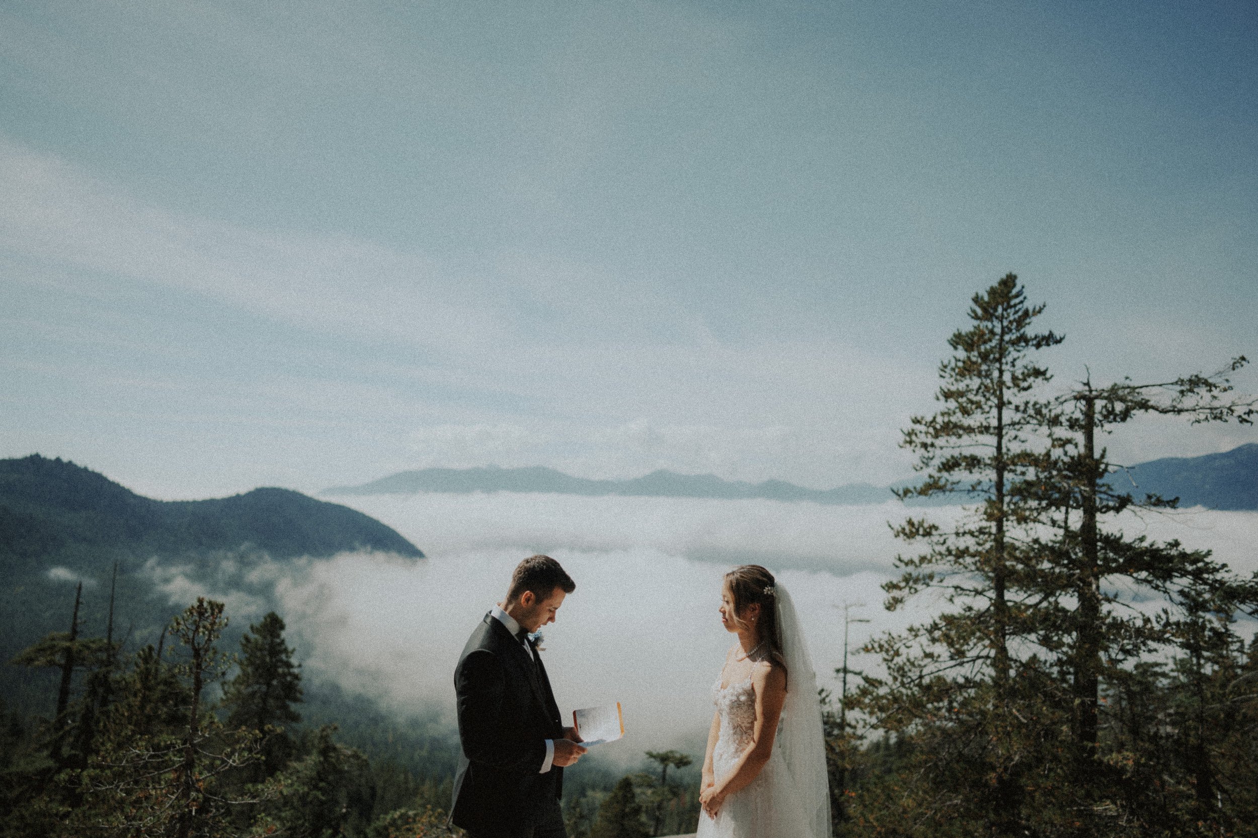 Couple exchanging rings against the mountain and cloud backdrop.