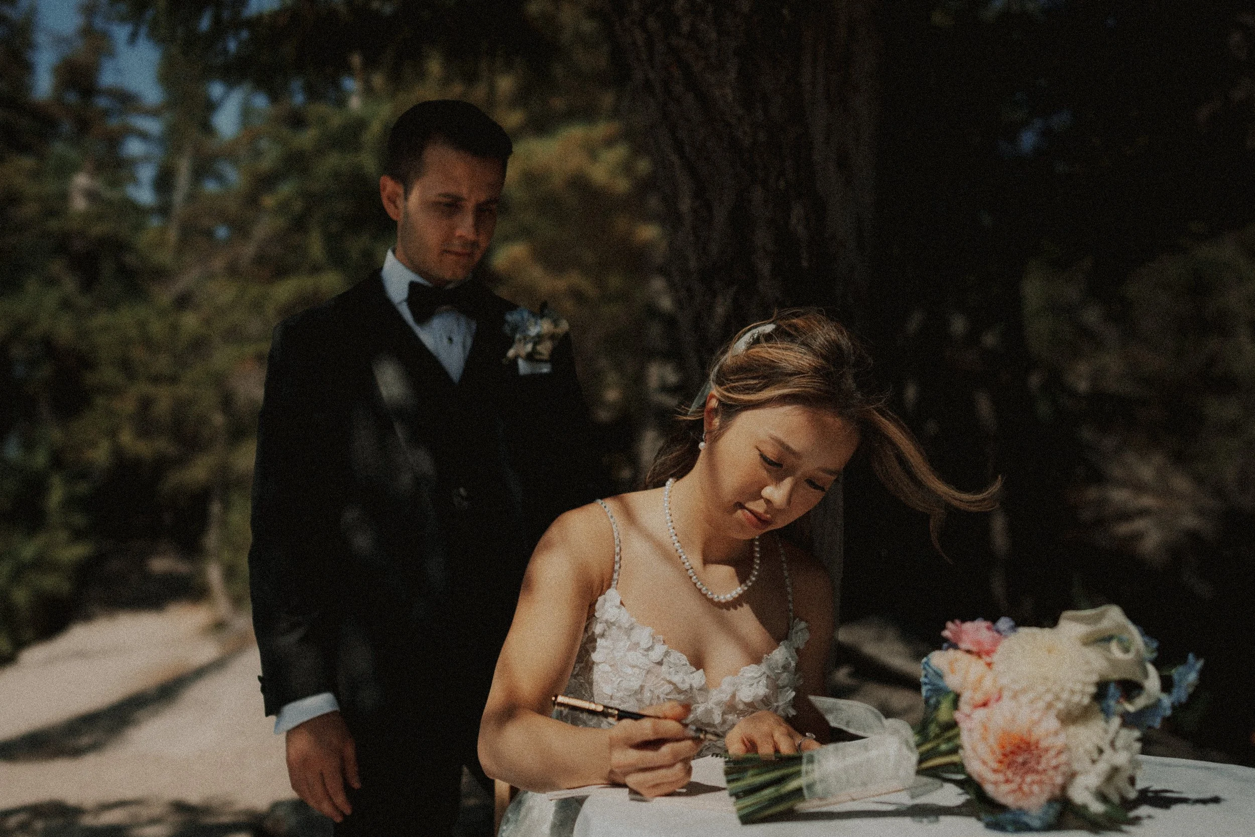 Bride signing wedding documents at an outdoor table beside the groom.