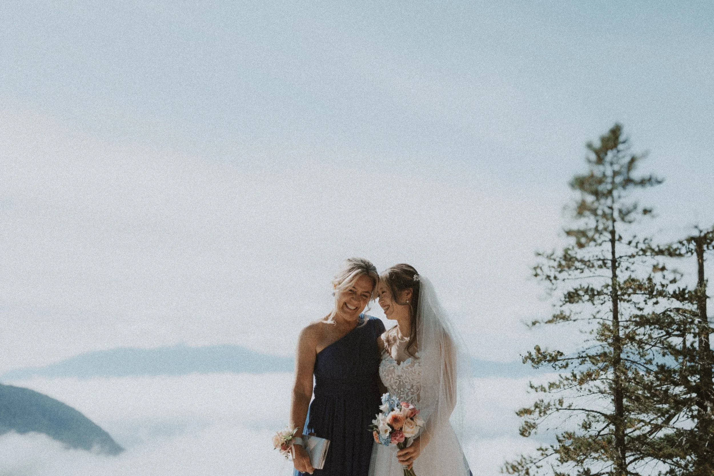 Bride leaning toward a guest as they pose together with the cloud-covered mountains behind.