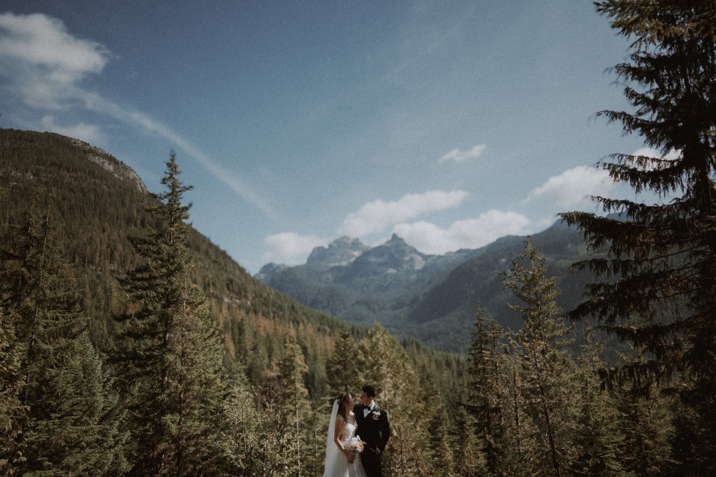 Newlyweds on a mountain trail beneath a wide blue sky and distant peaks.