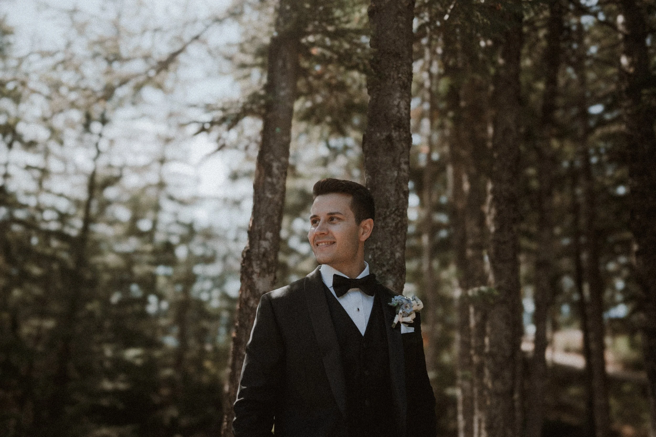 Groom smiling beside sunlit tree trunks in the mountain forest.