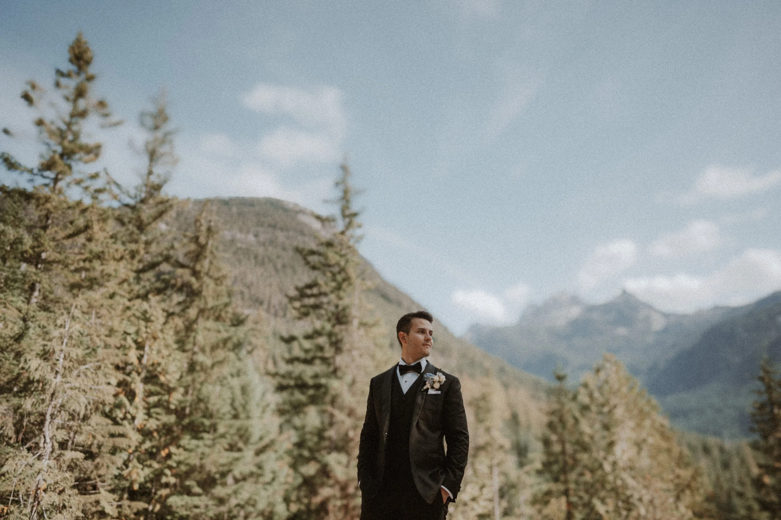 Groom standing alone with mountain peaks and forests behind him.