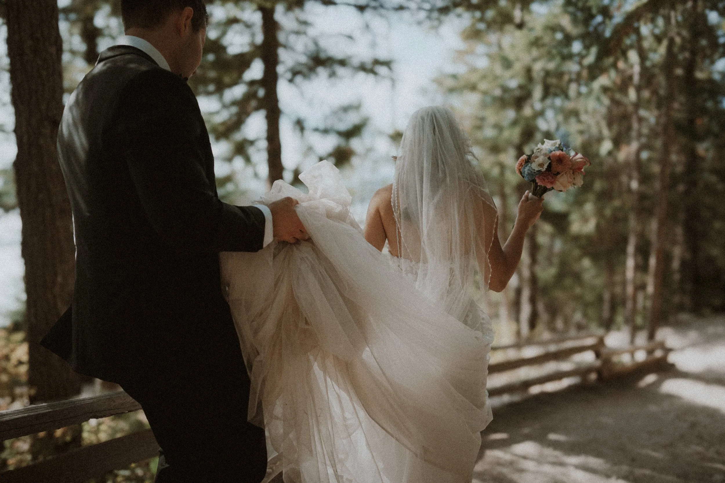 Groom lifting the bride's veil as she raises her bouquet beneath the trees.