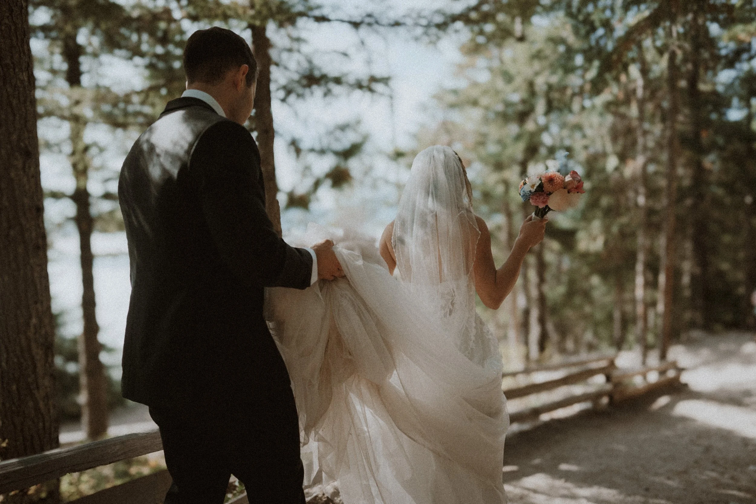 Couple walking through the forest with the bride's veil lifted behind her.