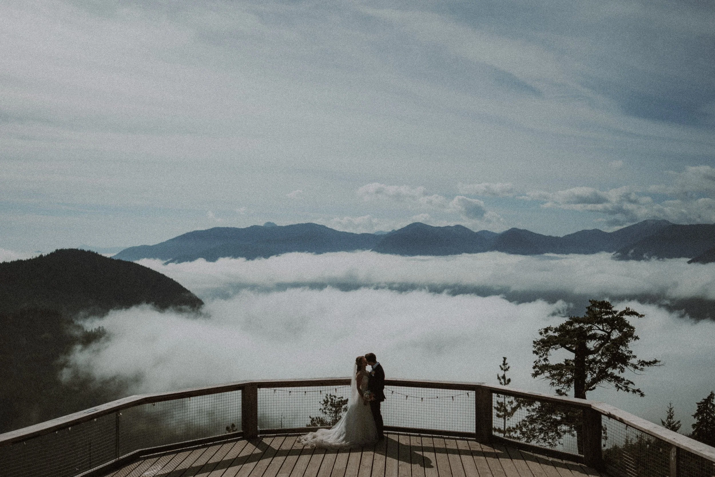 Couple standing on a timber viewing deck above a cloud-filled valley.