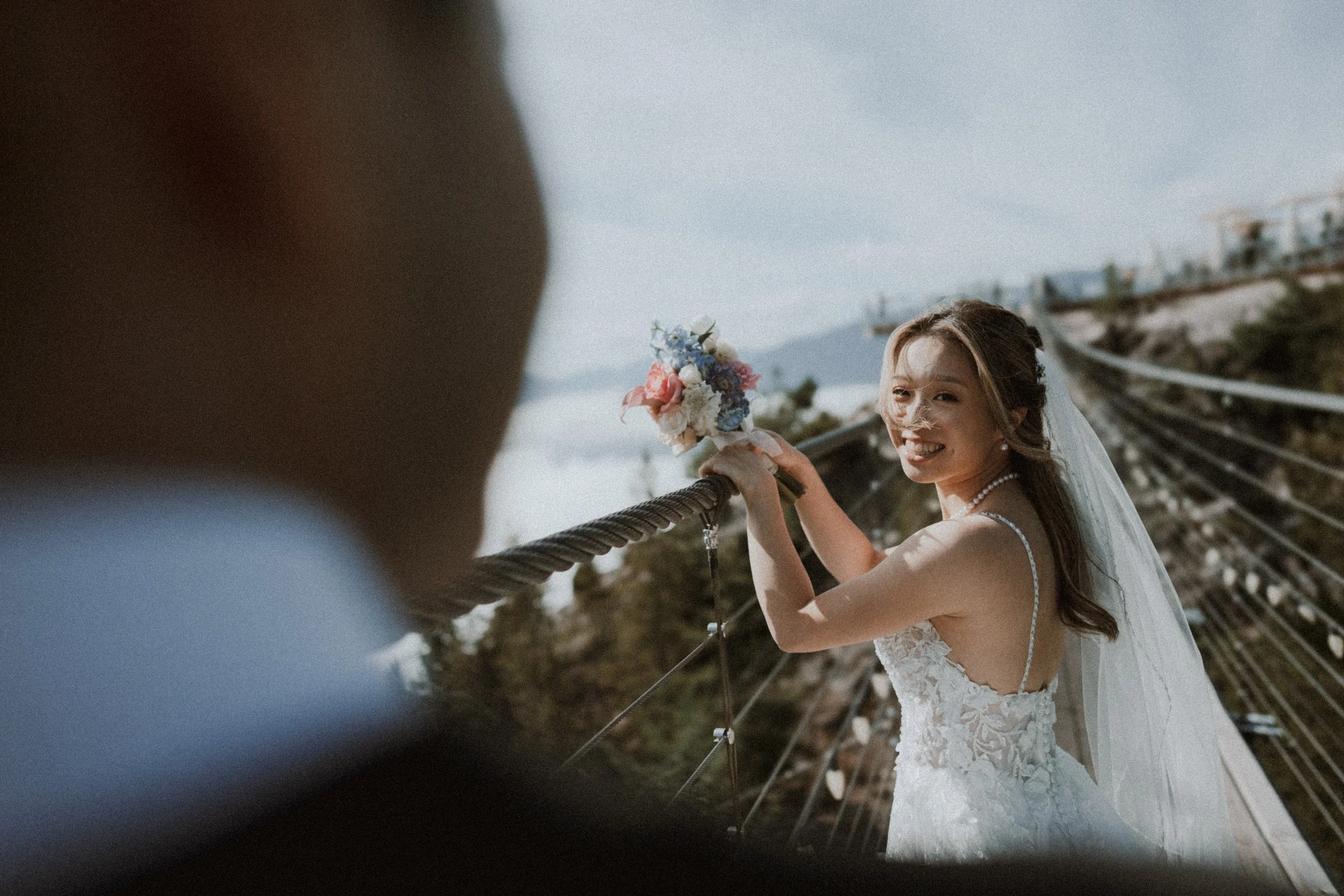 Bride smiling back at the groom while lifting her bouquet on the suspension bridge.