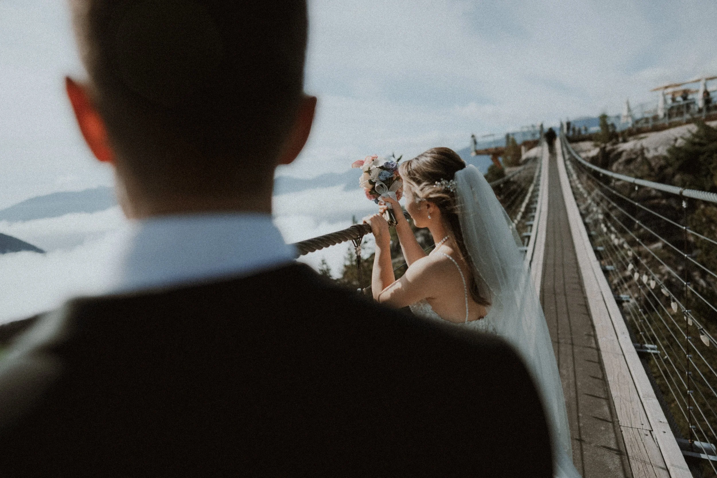View past the groom toward the bride on the mountain suspension bridge.