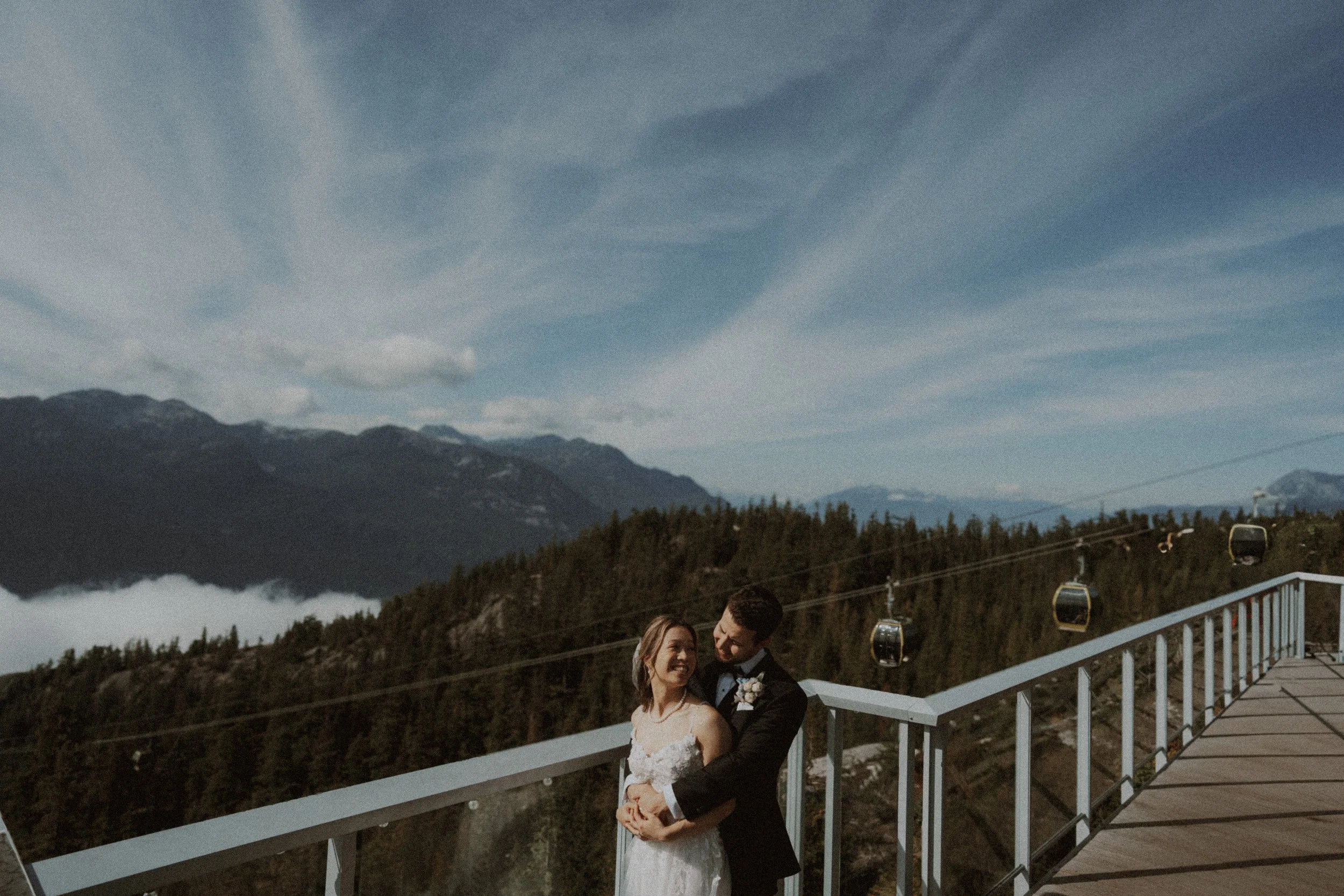 Couple embracing beside a railing with gondola cabins and mountains behind them.