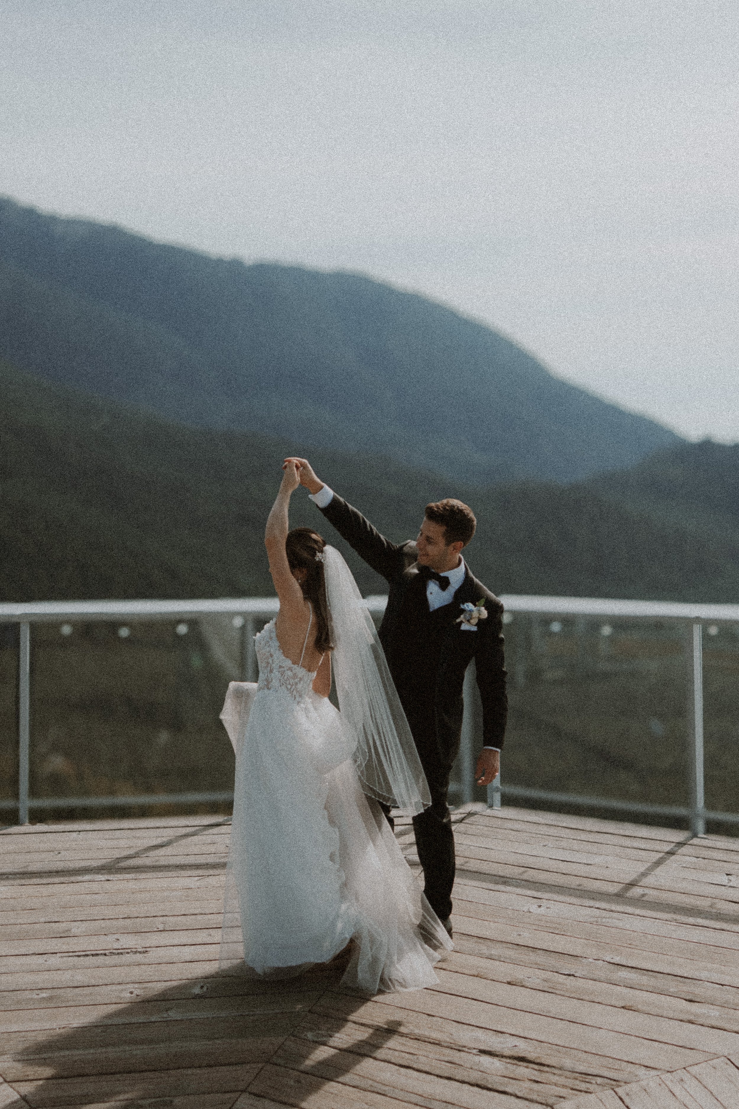 Bride raising her arm as the groom turns her on the mountain viewing deck.