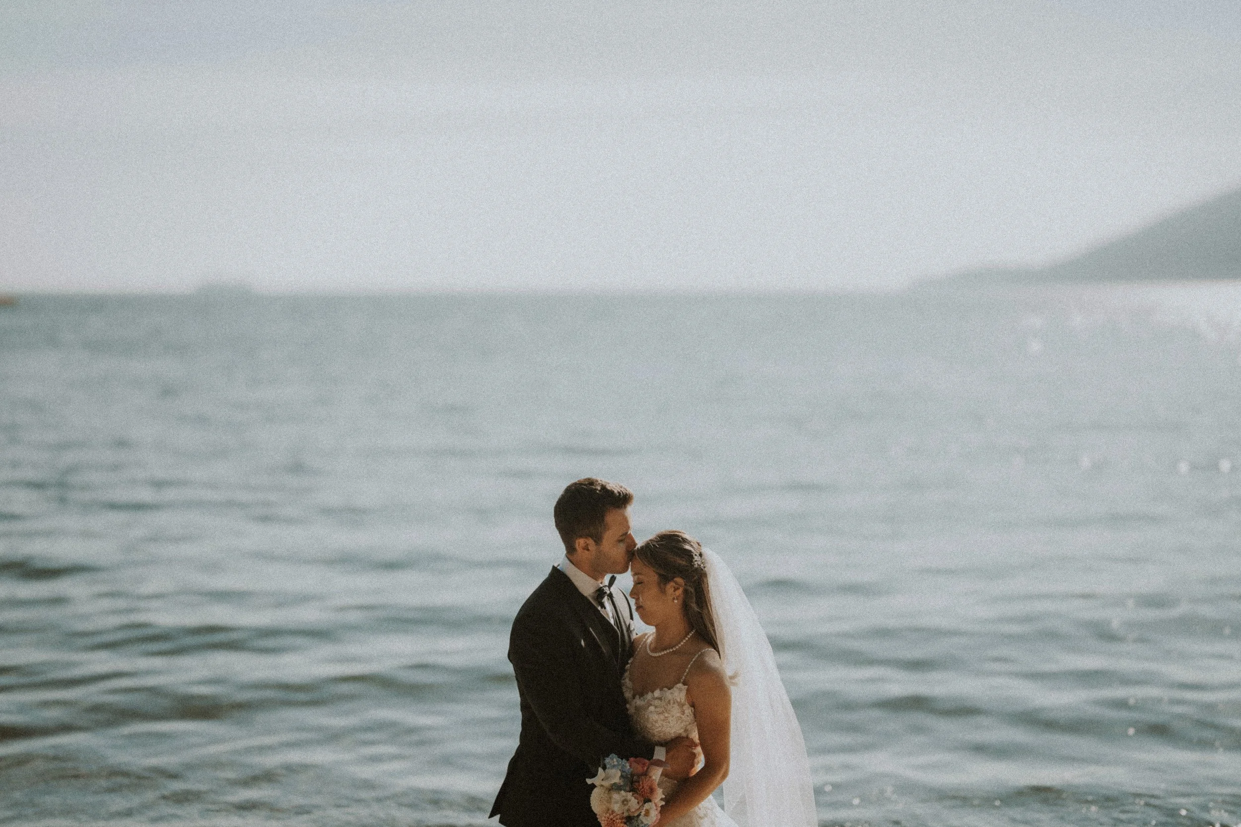 Couple sharing a quiet kiss with the coastal water behind them.