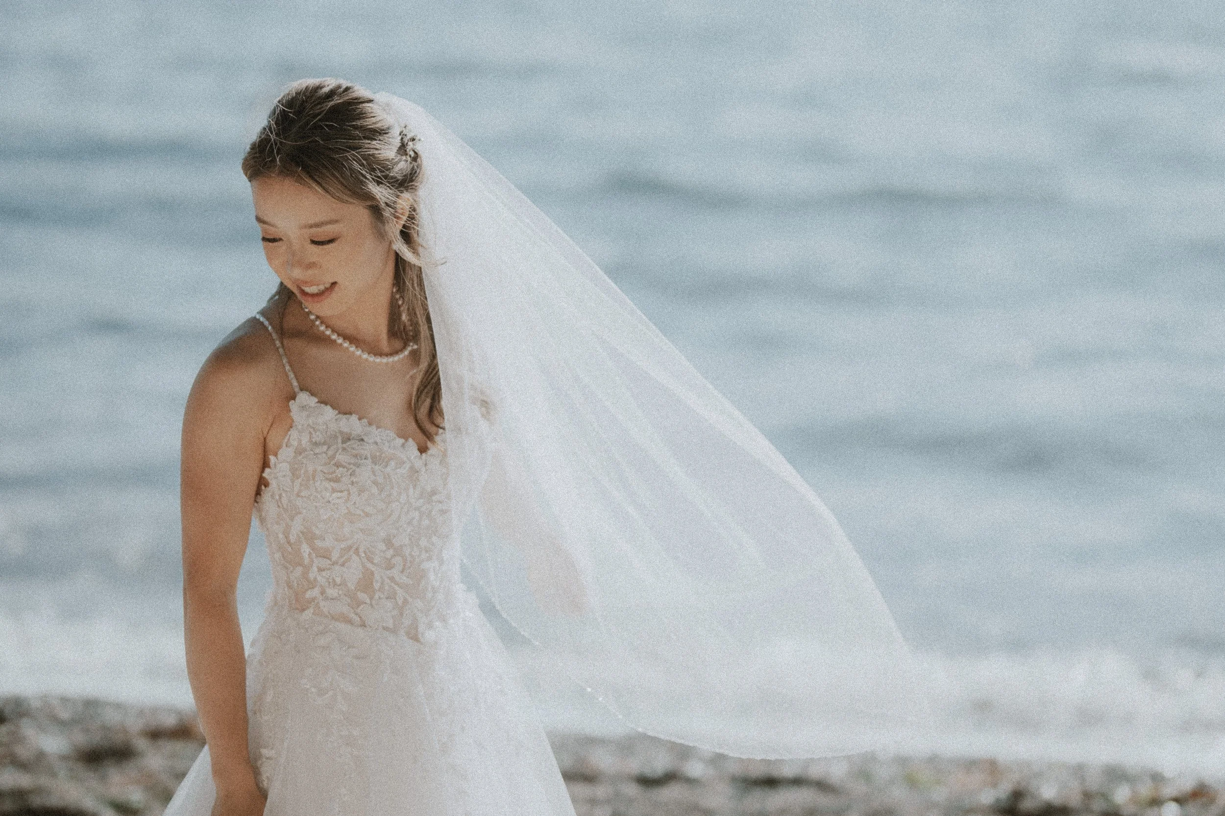 Bride looking down as her veil catches the breeze beside the water.