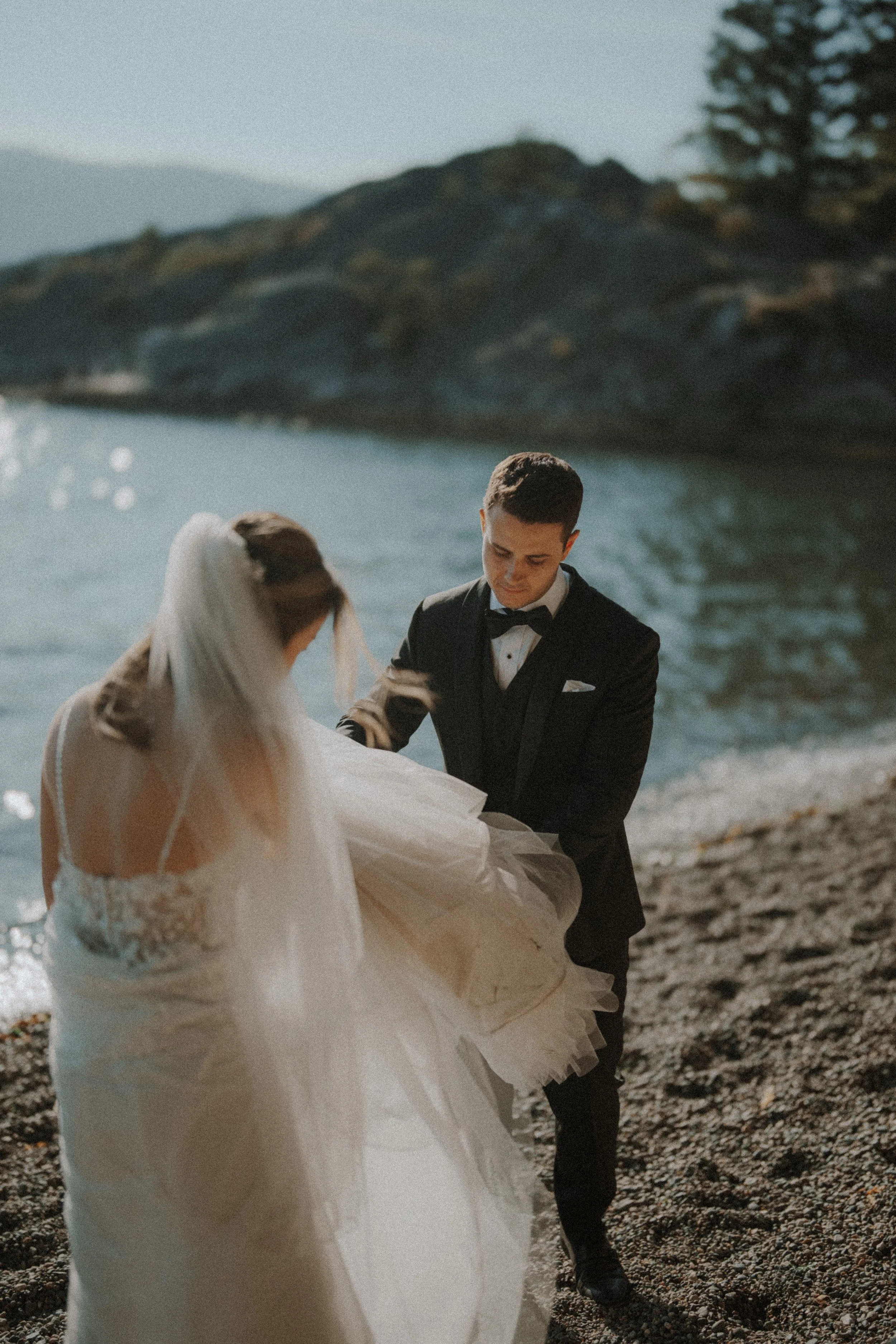 Groom helping hold the bride's gown above the pebbled shoreline.