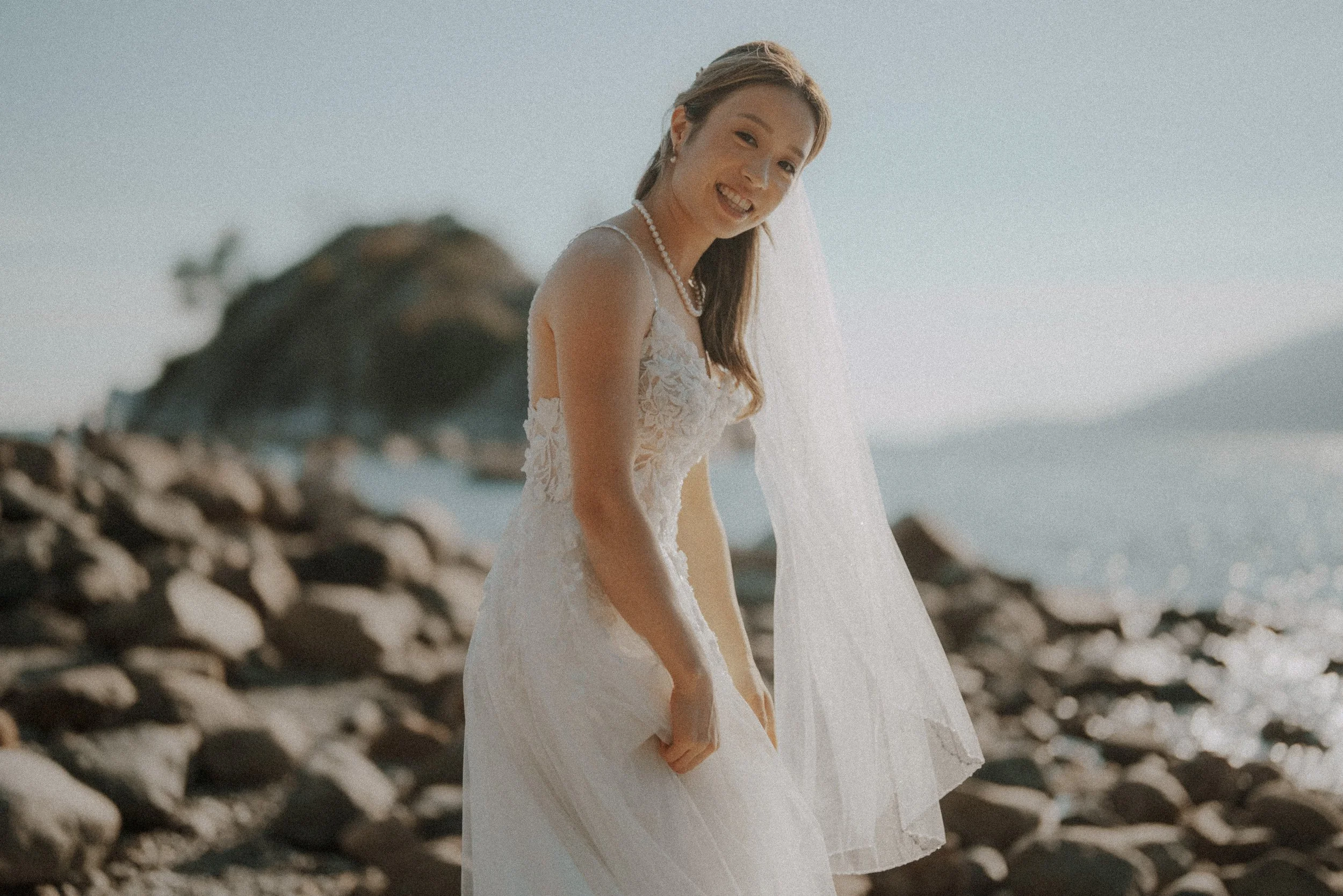 Bride smiling beside sunlit rocks and water at Whytecliff Park.