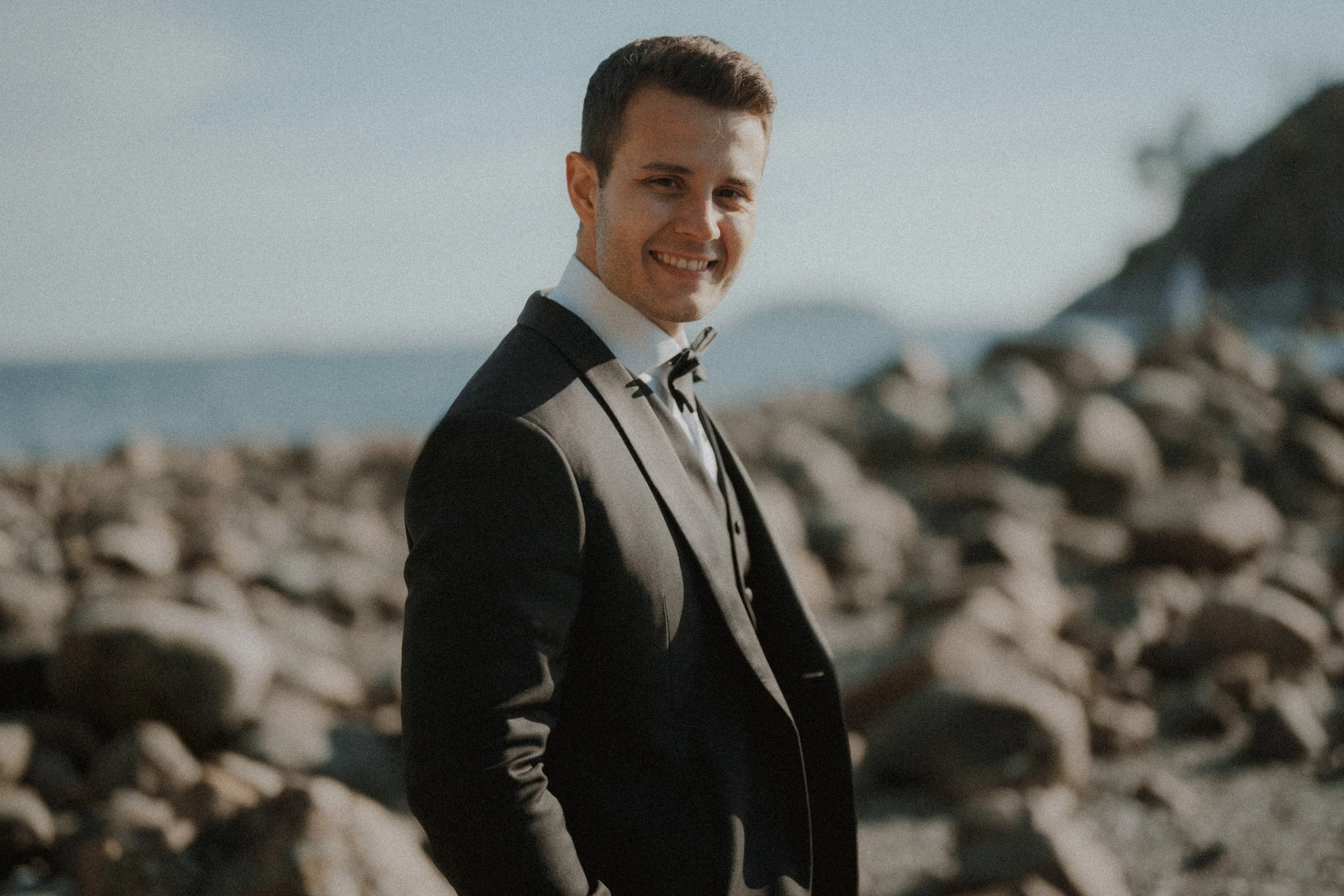Smiling groom in a black tuxedo beside sunlit coastal rocks.