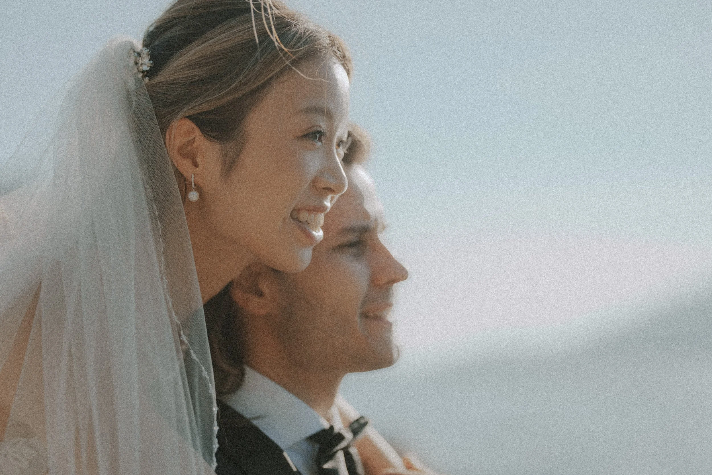 Bride resting her cheek beside the groom's head in soft coastal light.