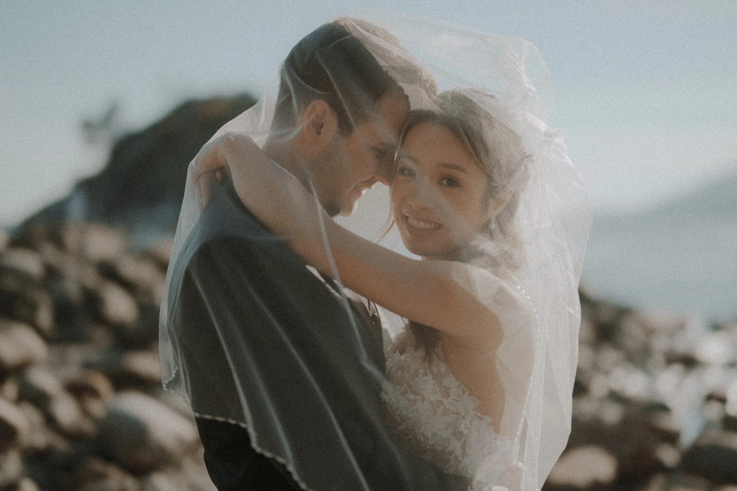 Bride smiling toward the camera while embracing the groom beneath her veil.
