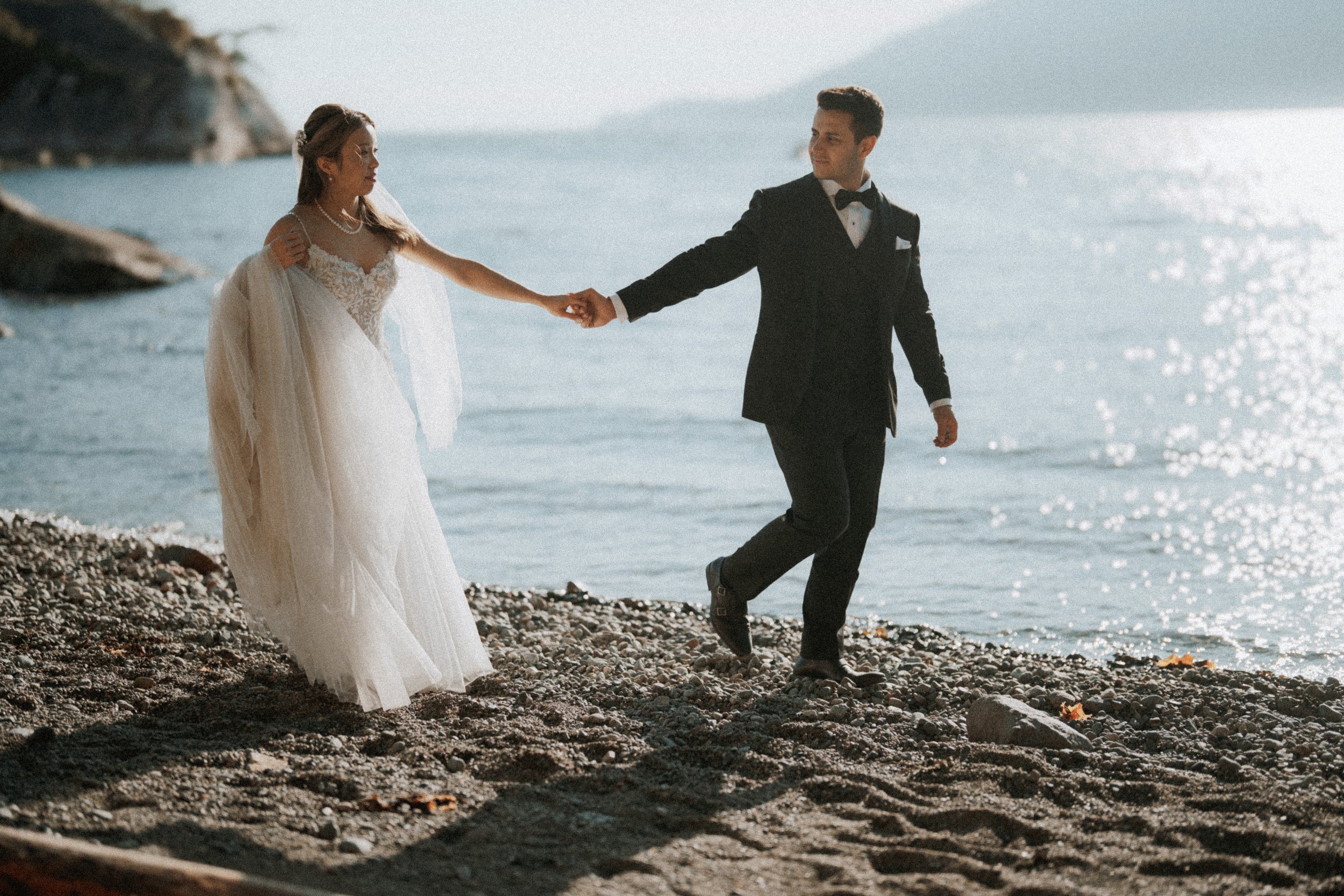 Newlyweds holding hands and stepping across the pebbled beach.