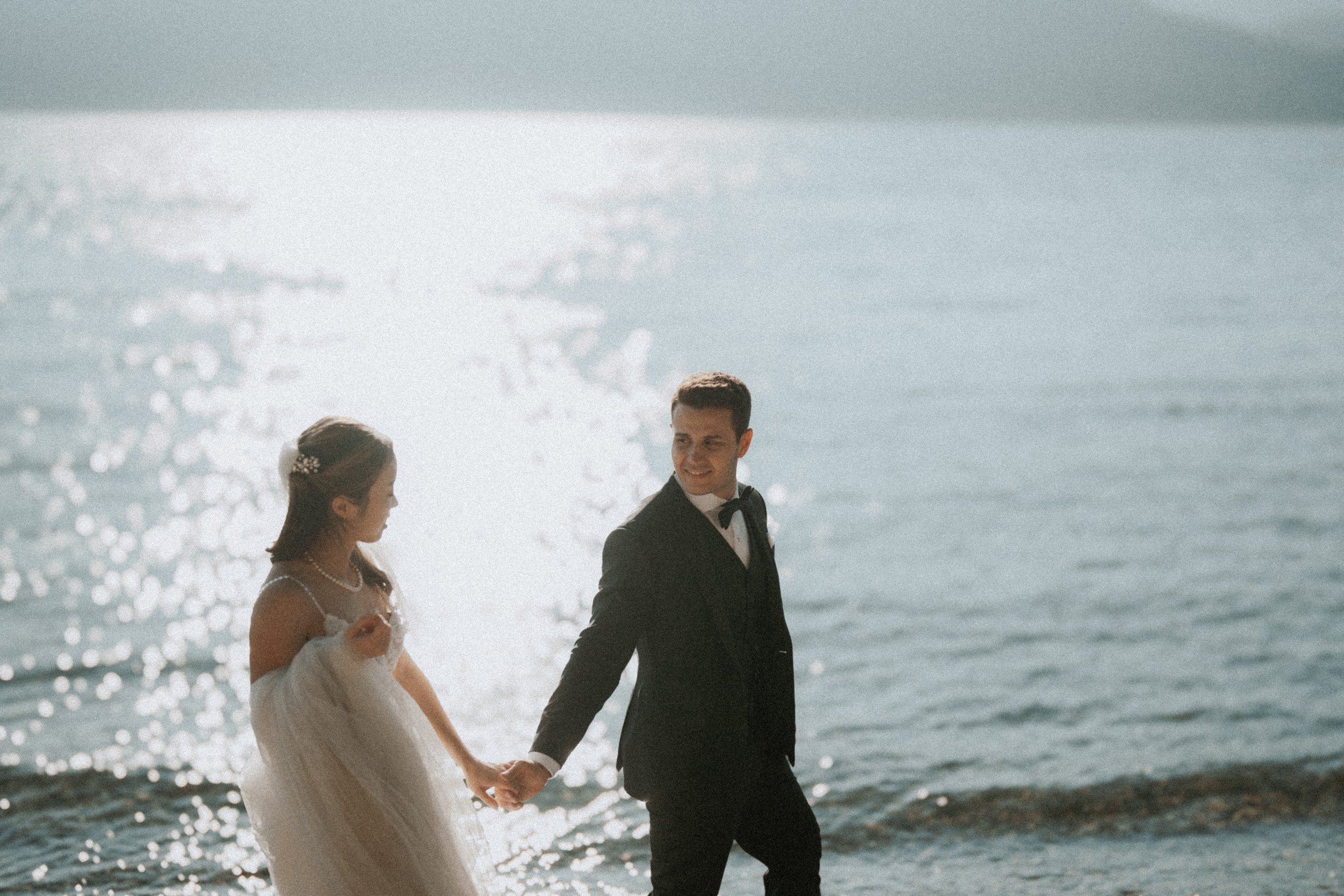 Bride and groom walking beside sparkling water on the pebbled beach.