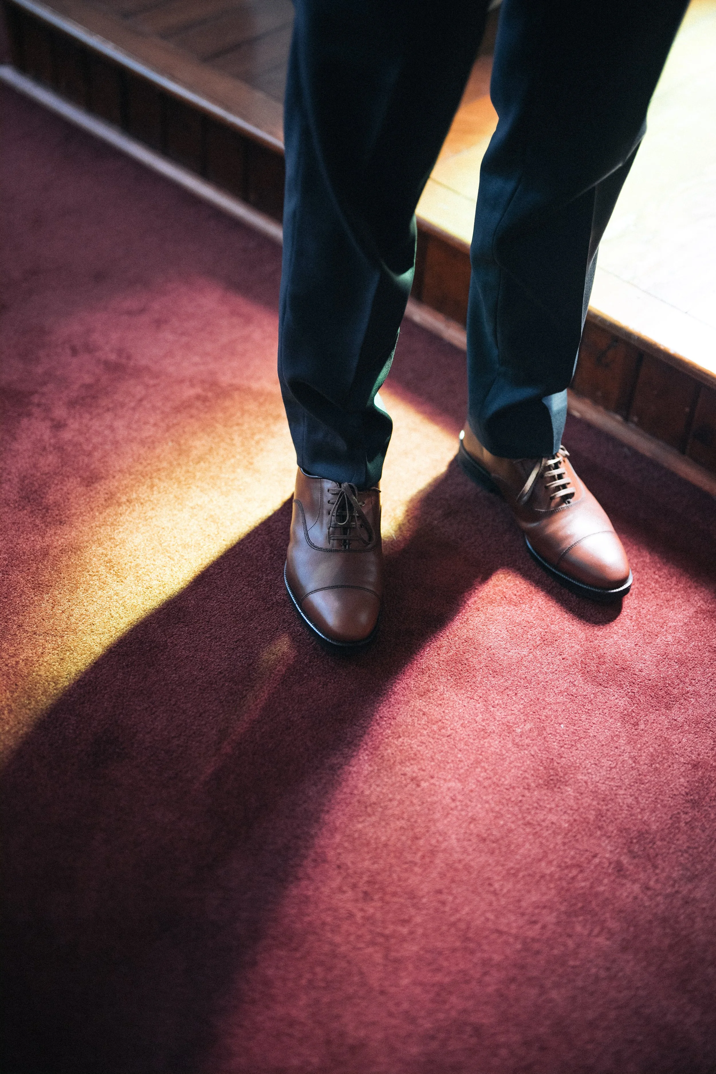 Groom's polished brown shoes and dark trousers on a sunlit chapel carpet.