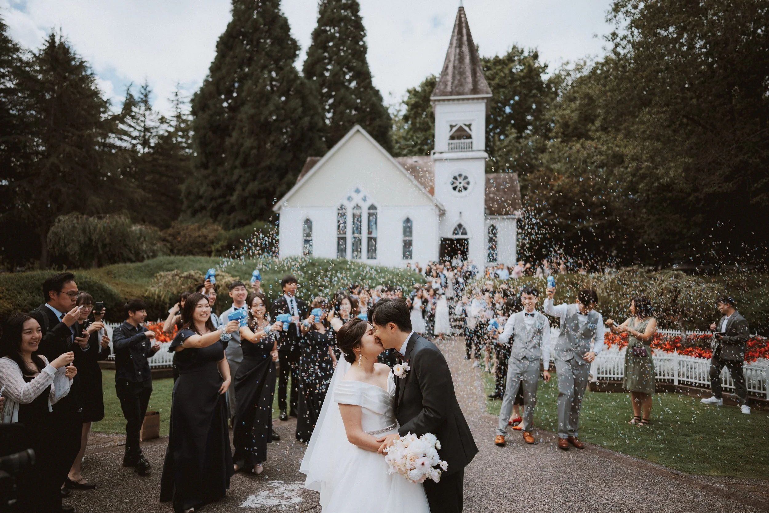 Wide view of the newlyweds kissing among guests and bubbles outside the chapel.
